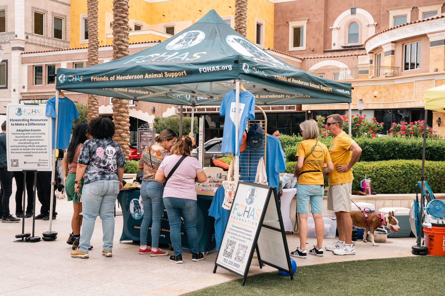 People gathered around outdoor vendor tent at community event with palm trees in background.