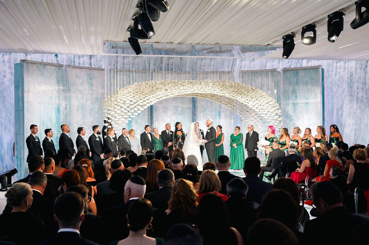 Wedding ceremony scene with floral arch and gathering of guests in an elegant white and blue themed venue.