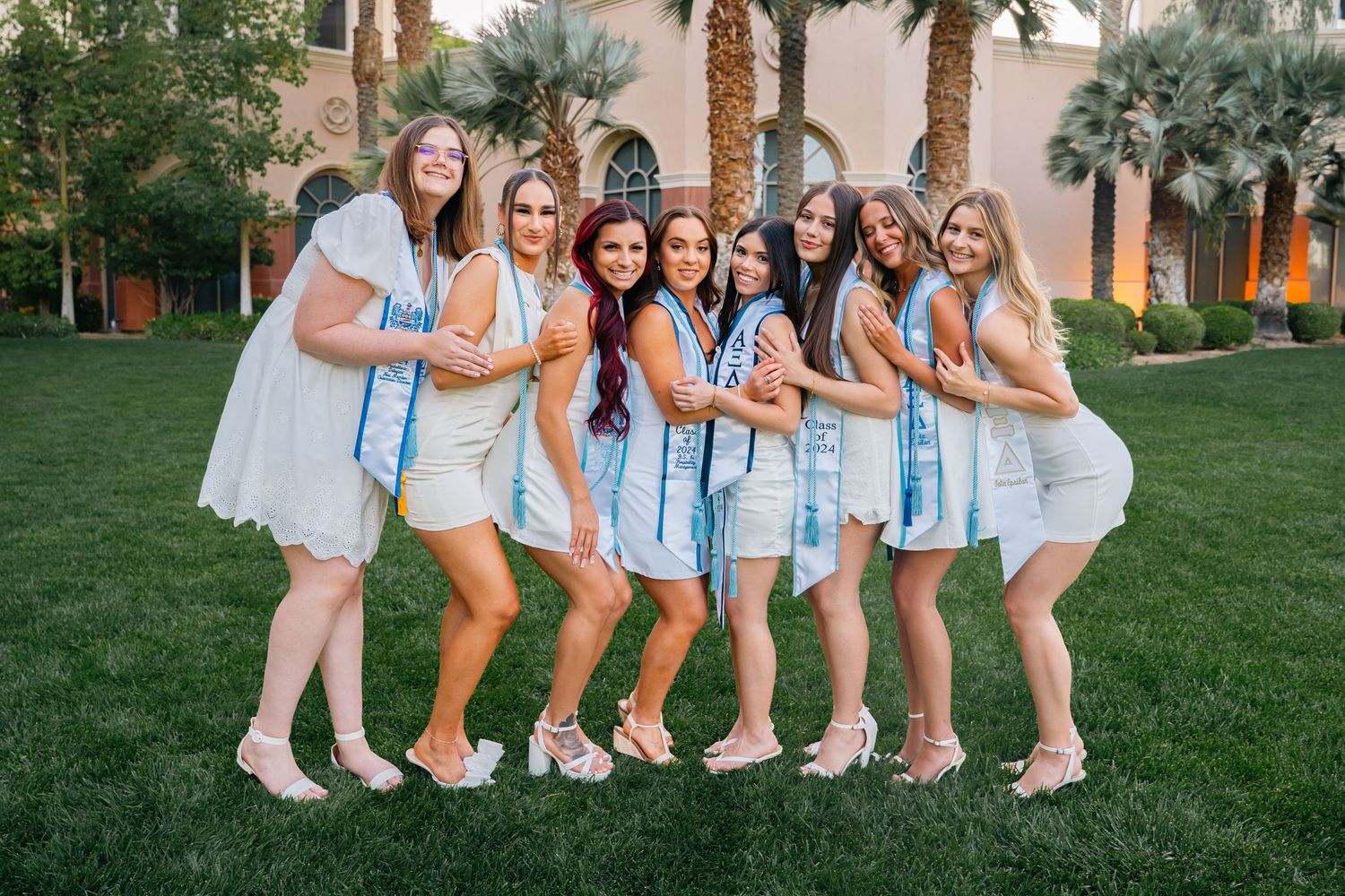 Group of sorority sisters in white dresses pose together with matching light blue stoles on campus lawn.
