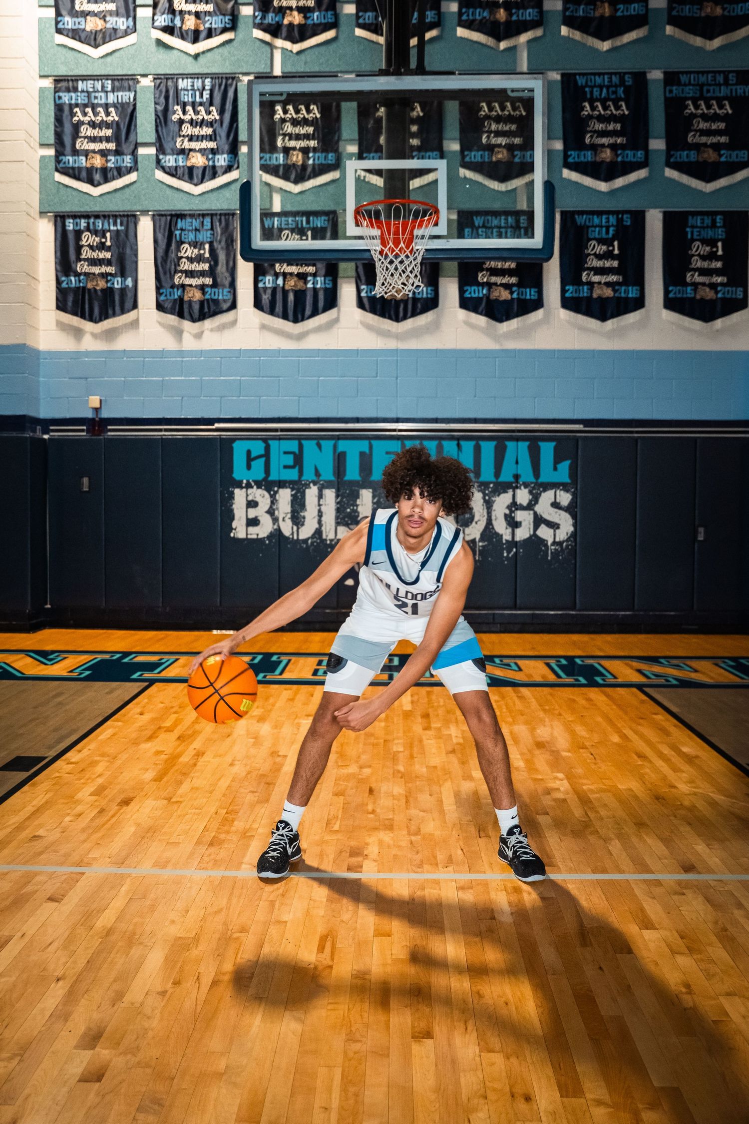 Basketball player in white uniform dribbles low on wooden court with championship banners displayed.
