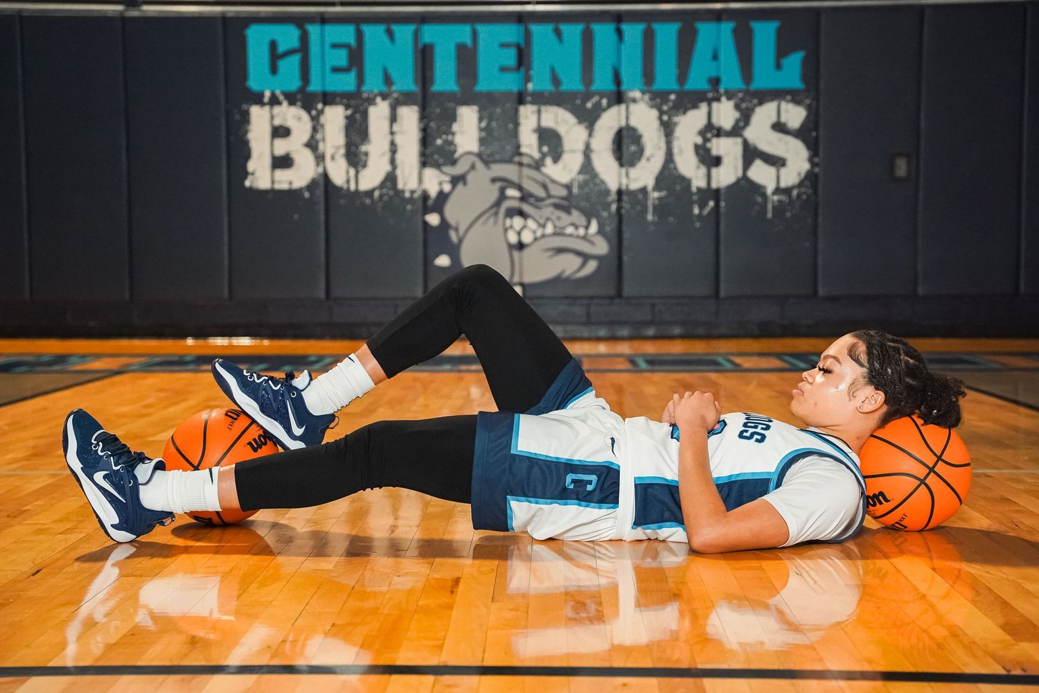 Basketball player lounges on gym floor surrounded by basketballs in Centennial uniform.
