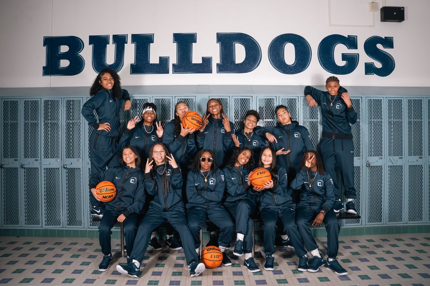 Team photo of Bulldogs basketball squad in navy warmups against school wall.