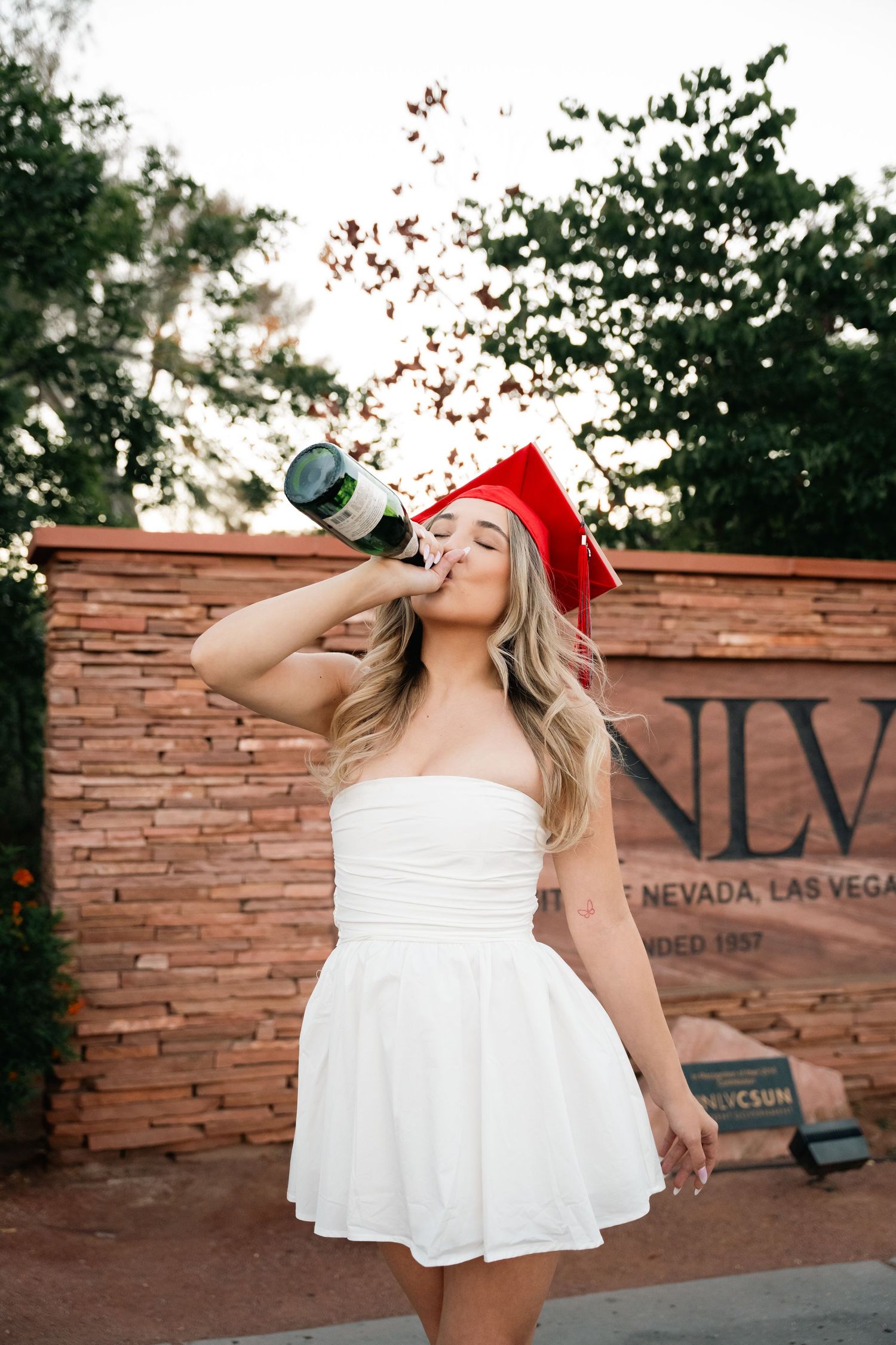 Graduate celebrates with champagne in front of UNLV stone wall sign.