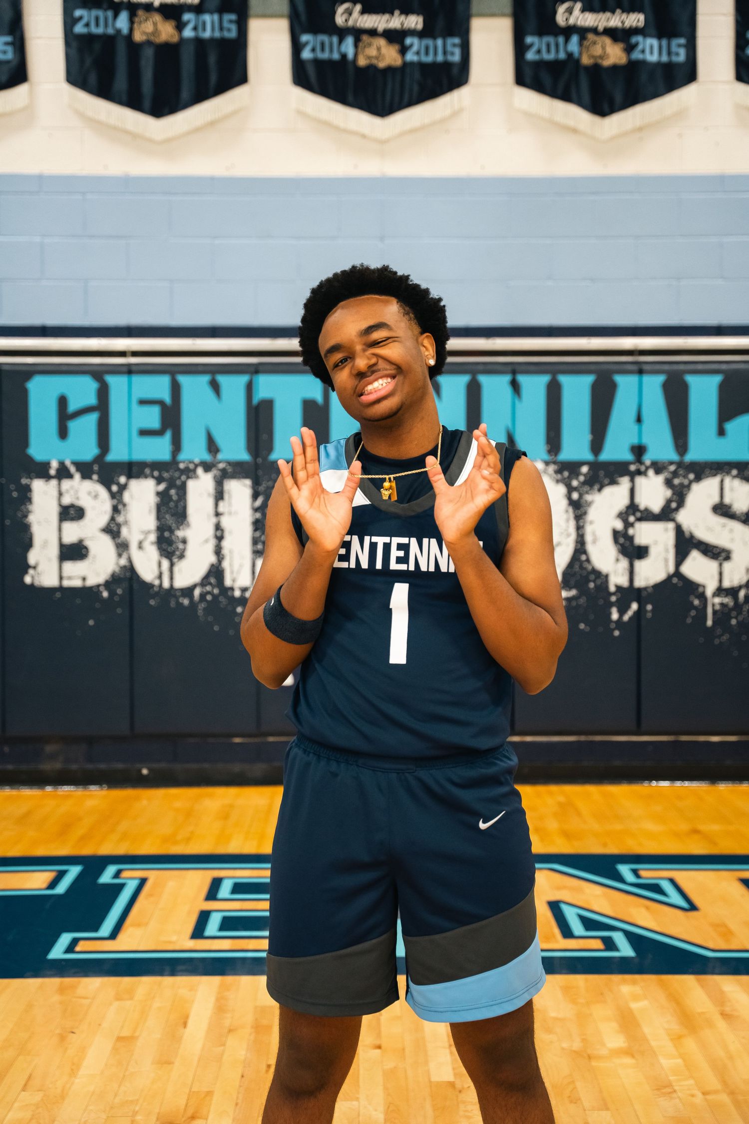 Basketball player in navy uniform poses energetically on court with Centennial Bulldogs signage.