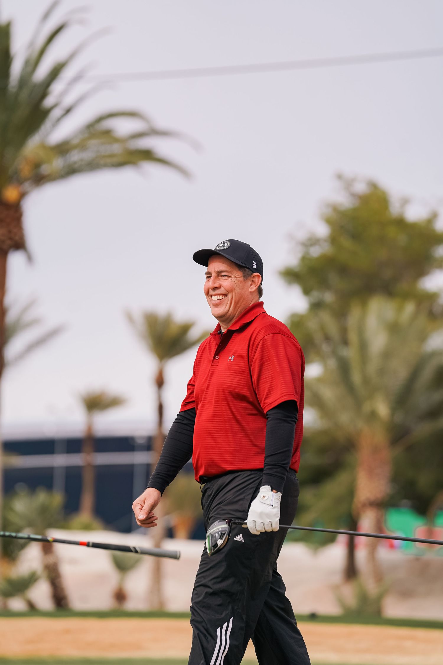Smiling golfer in red polo and black layers walking on desert golf course with palm trees.