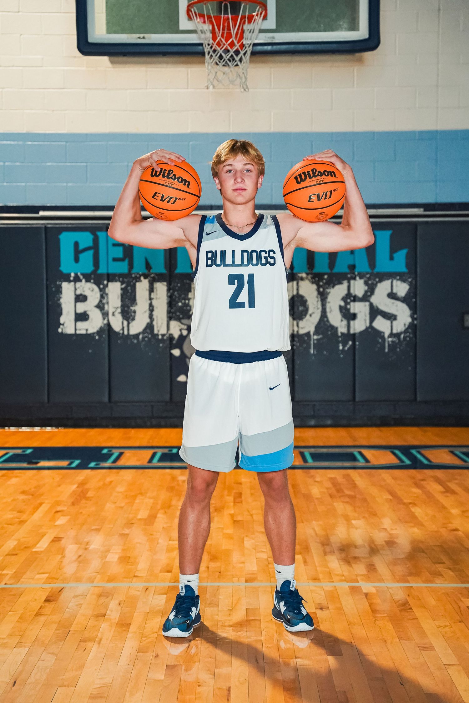 Basketball player in Bulldogs uniform holding two basketballs overhead in gym.