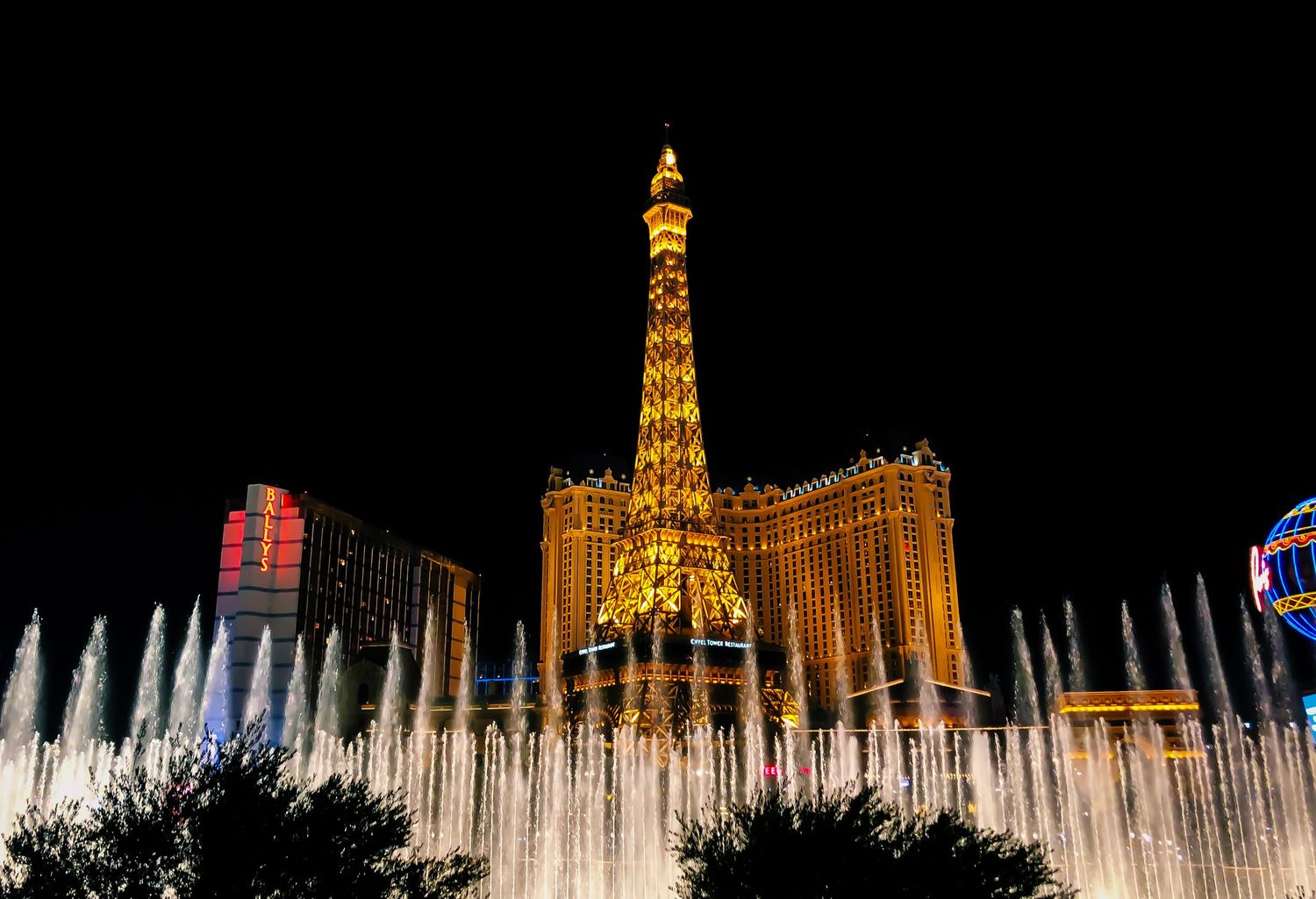 Bellagio fountains performing in front of illuminated Paris Las Vegas hotel at night.