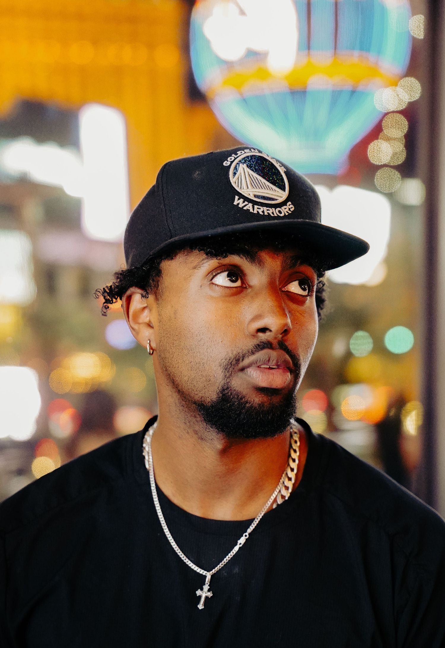 Person in black Golden State Warriors cap and necklace with cross pendant looking to the side at night.