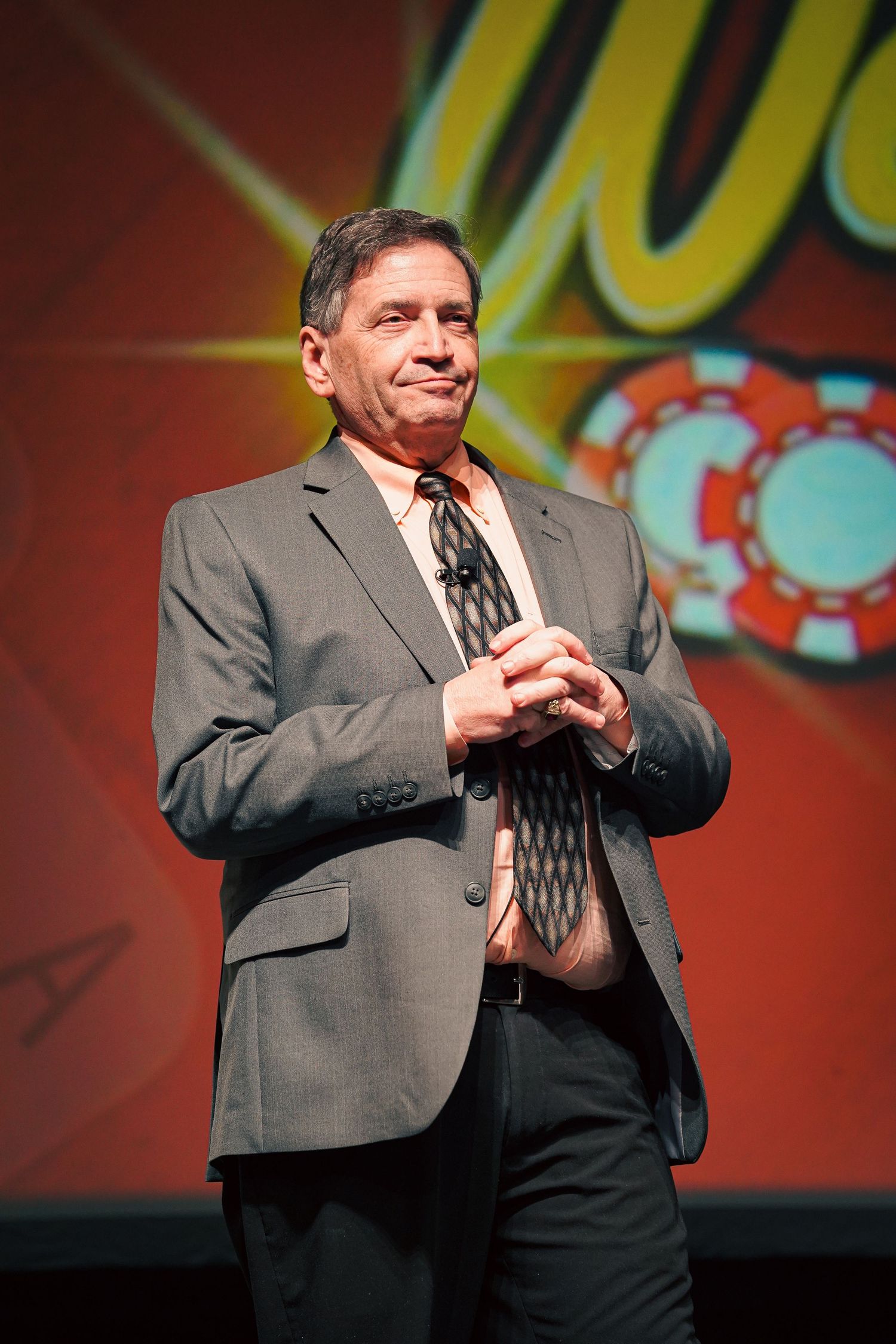 Man in gray suit gesturing on stage with red backdrop at Vegas event.
