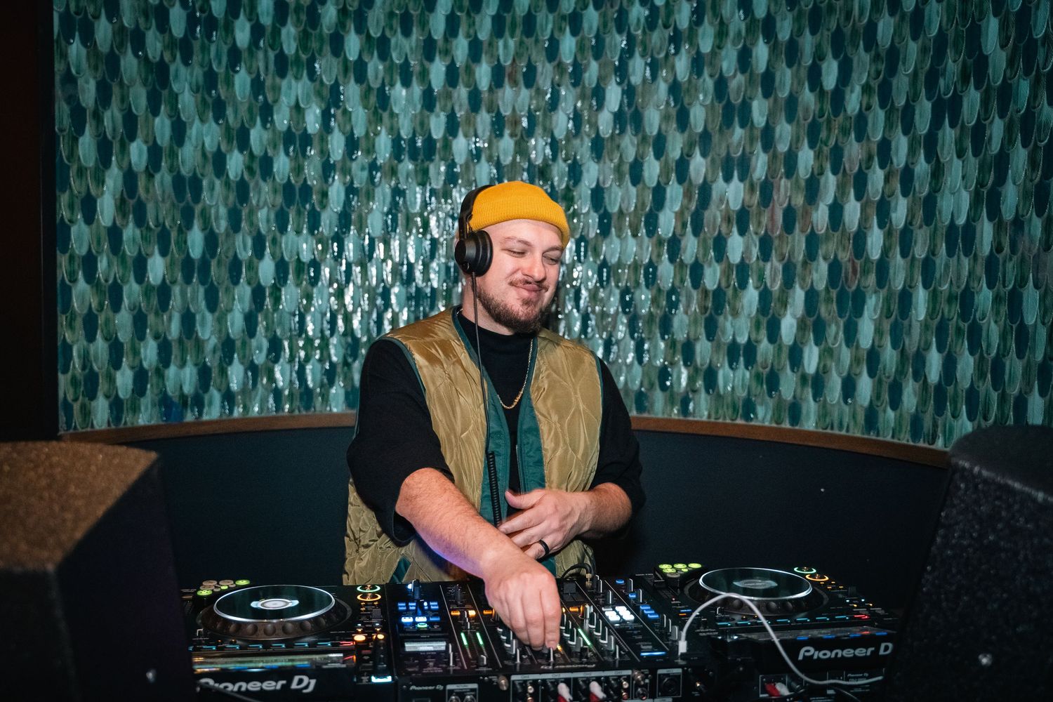 A DJ performs at a booth with turntables against a patterned teal background.