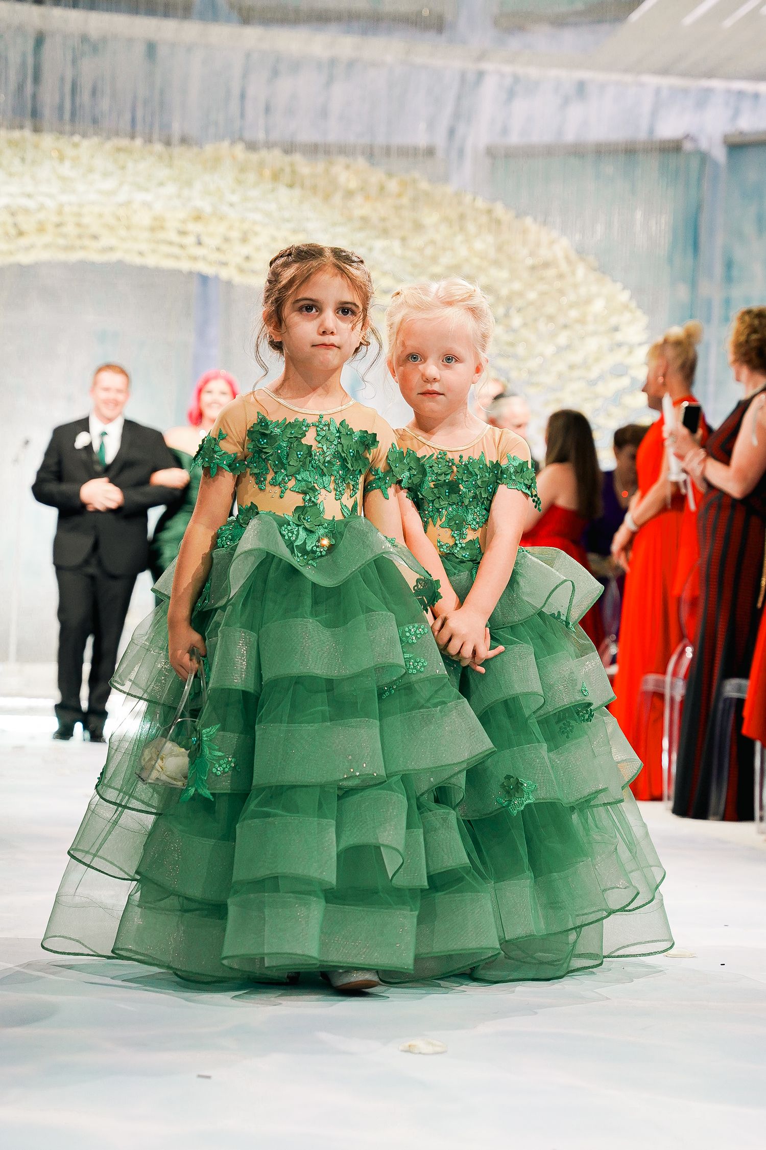 Two flower girls in matching emerald green tiered ballgowns hold hands walking down the aisle.