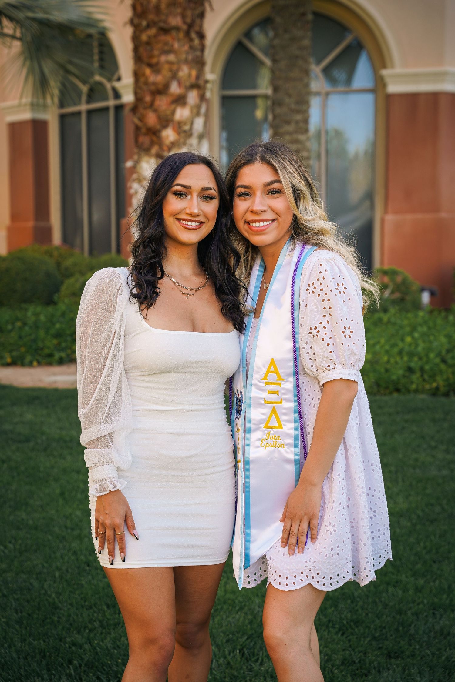 Two graduates in white dresses pose together with decorated sashes on resort lawn.