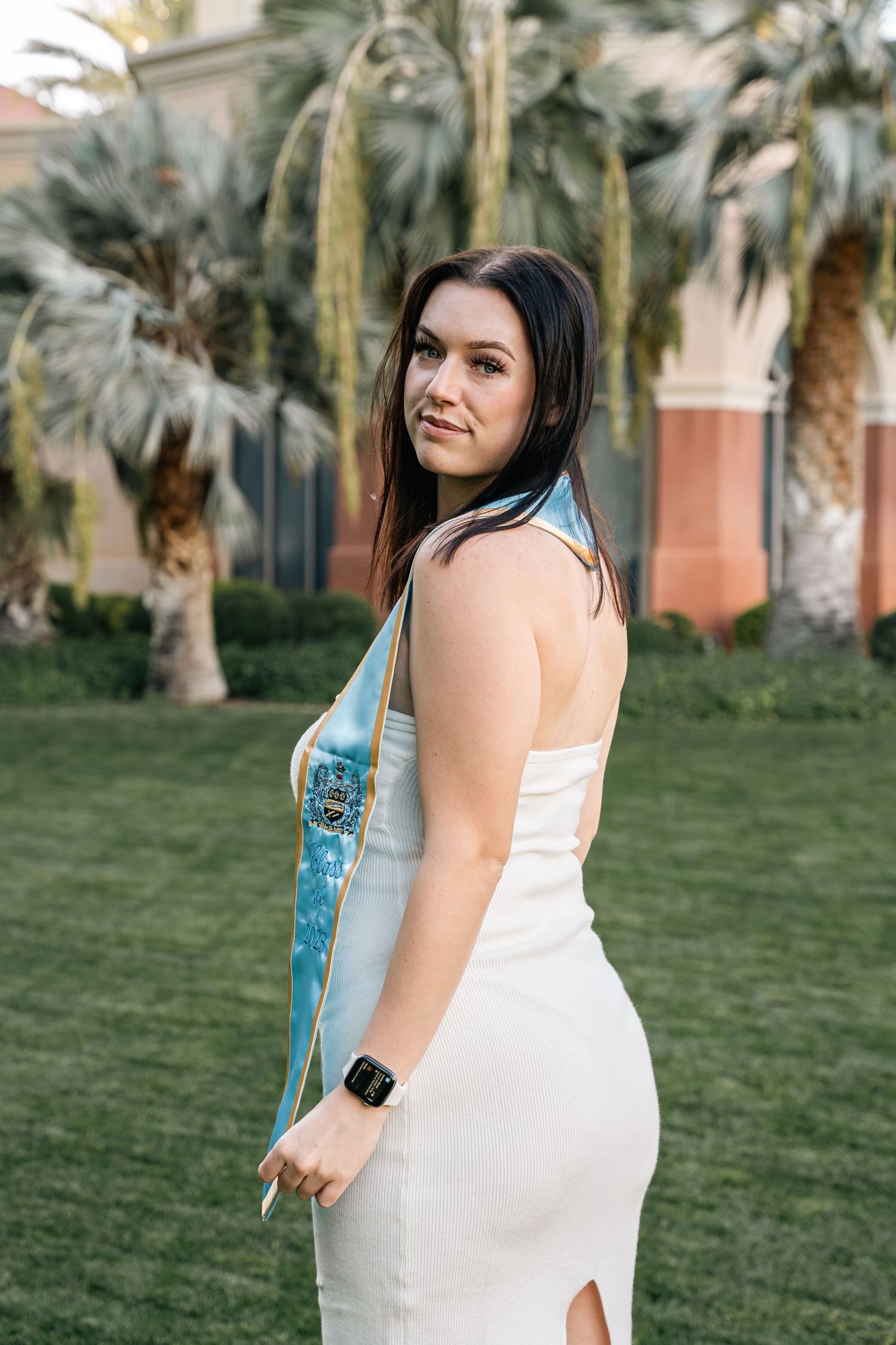White dress and blue sash outfit shown against tropical landscaping and Mediterranean architecture.