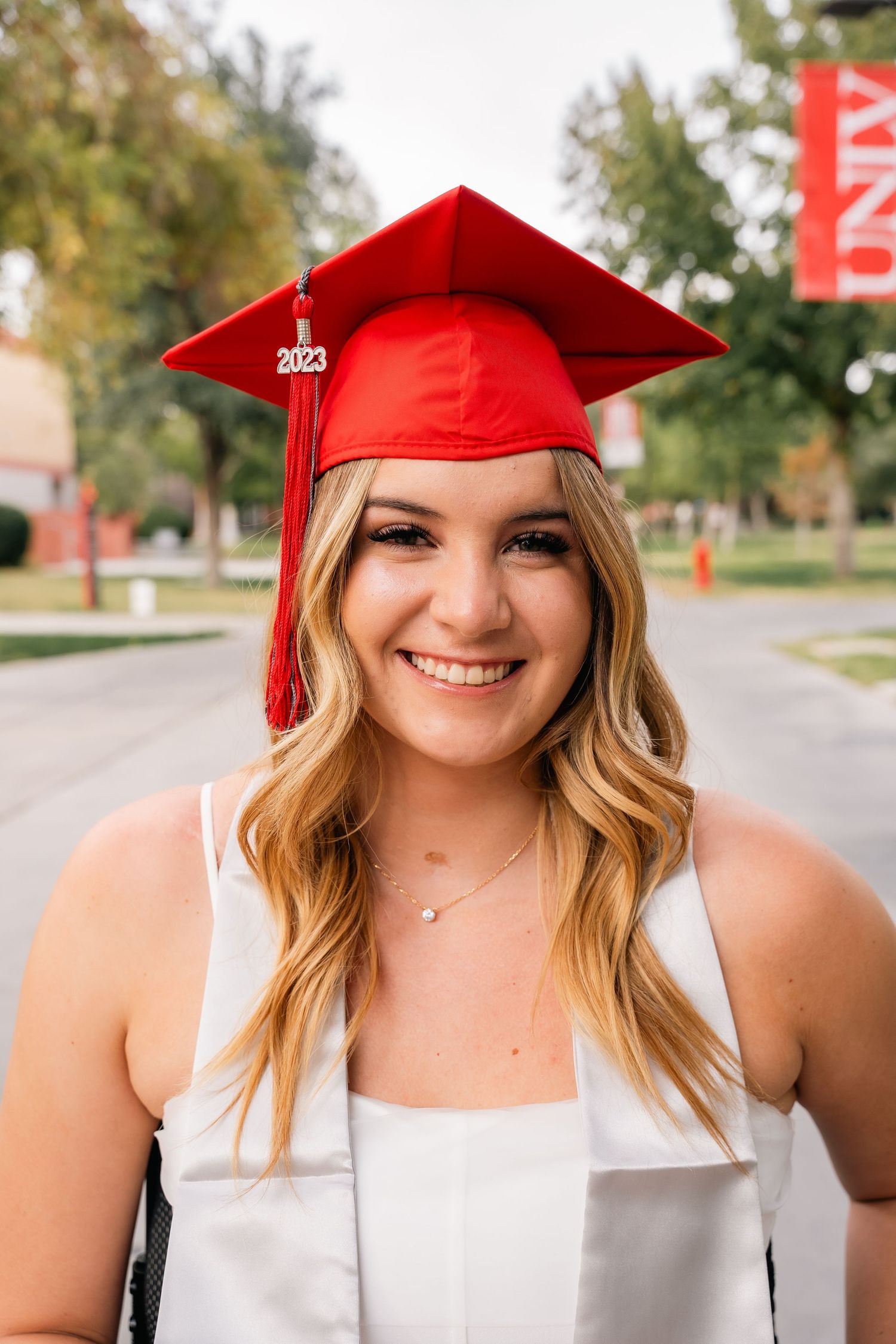 Graduate wearing 2023 red cap and white dress smiling outdoors on university campus.