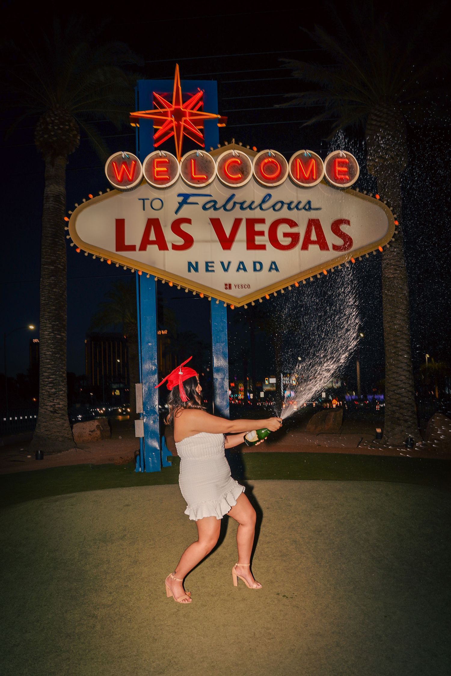 Woman popping champagne in front of iconic Las Vegas welcome sign at night.