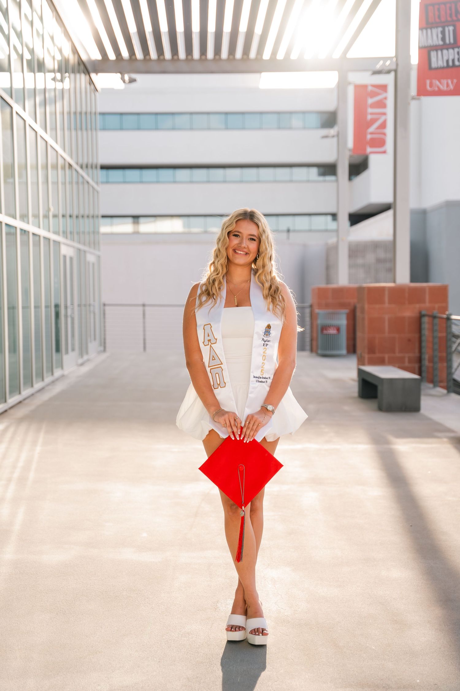 Graduate holds red cap while posing in white dress in modern university building hallway.