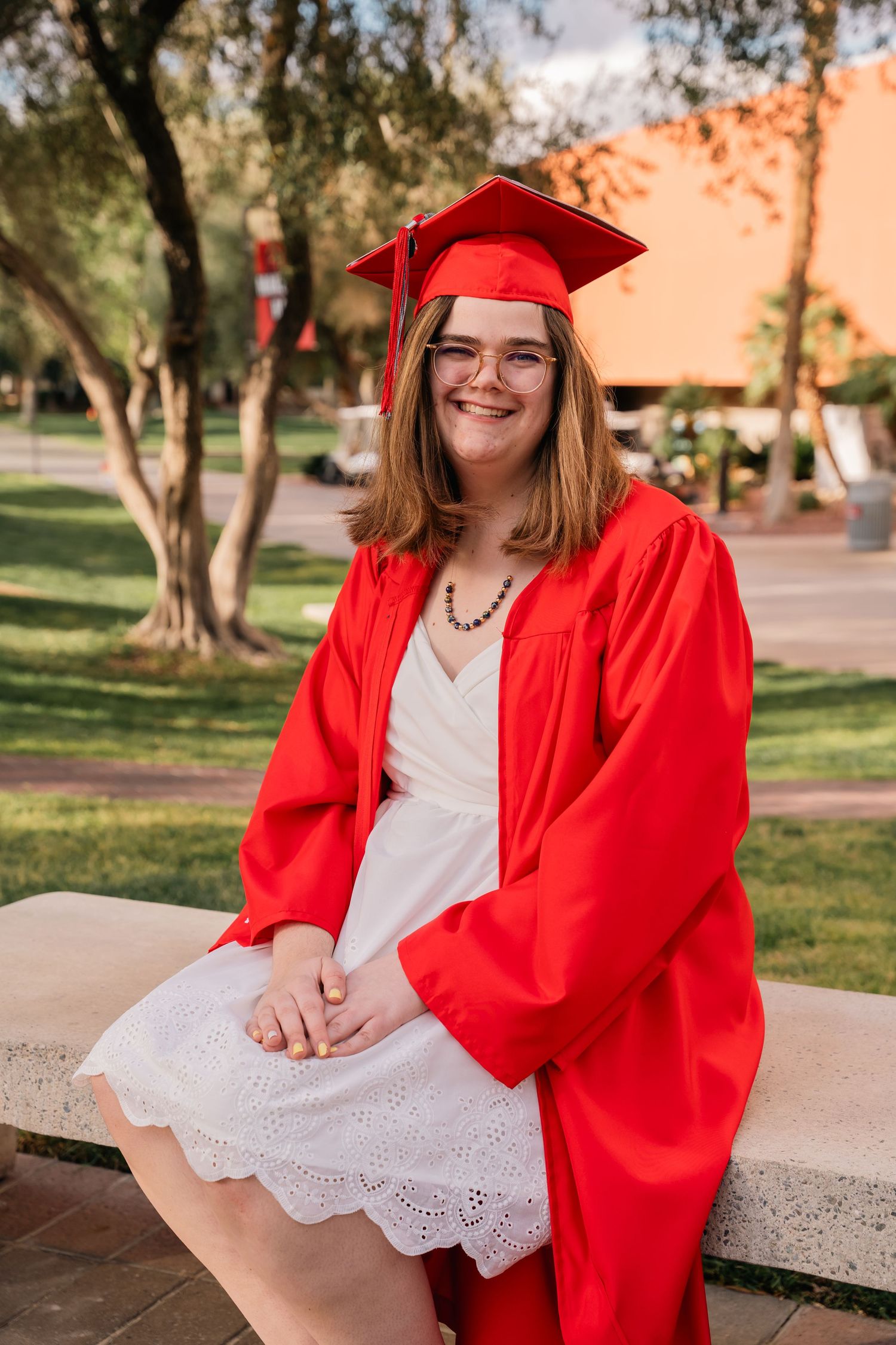 Graduate in red cap and gown sits outdoors on campus.