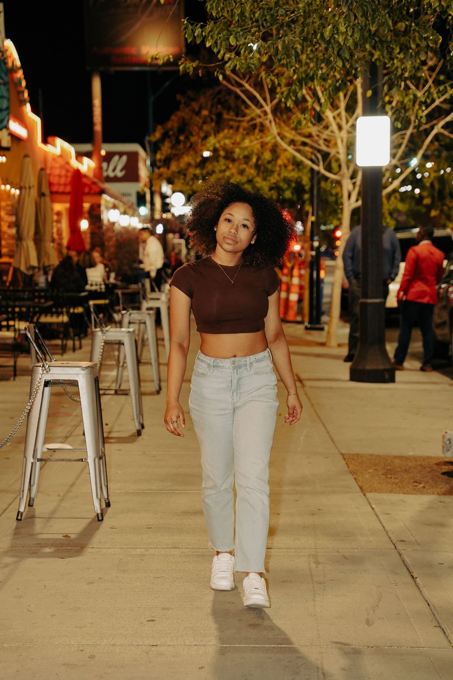 Stylish woman in crop top and jeans walks along urban sidewalk at night near restaurants.
