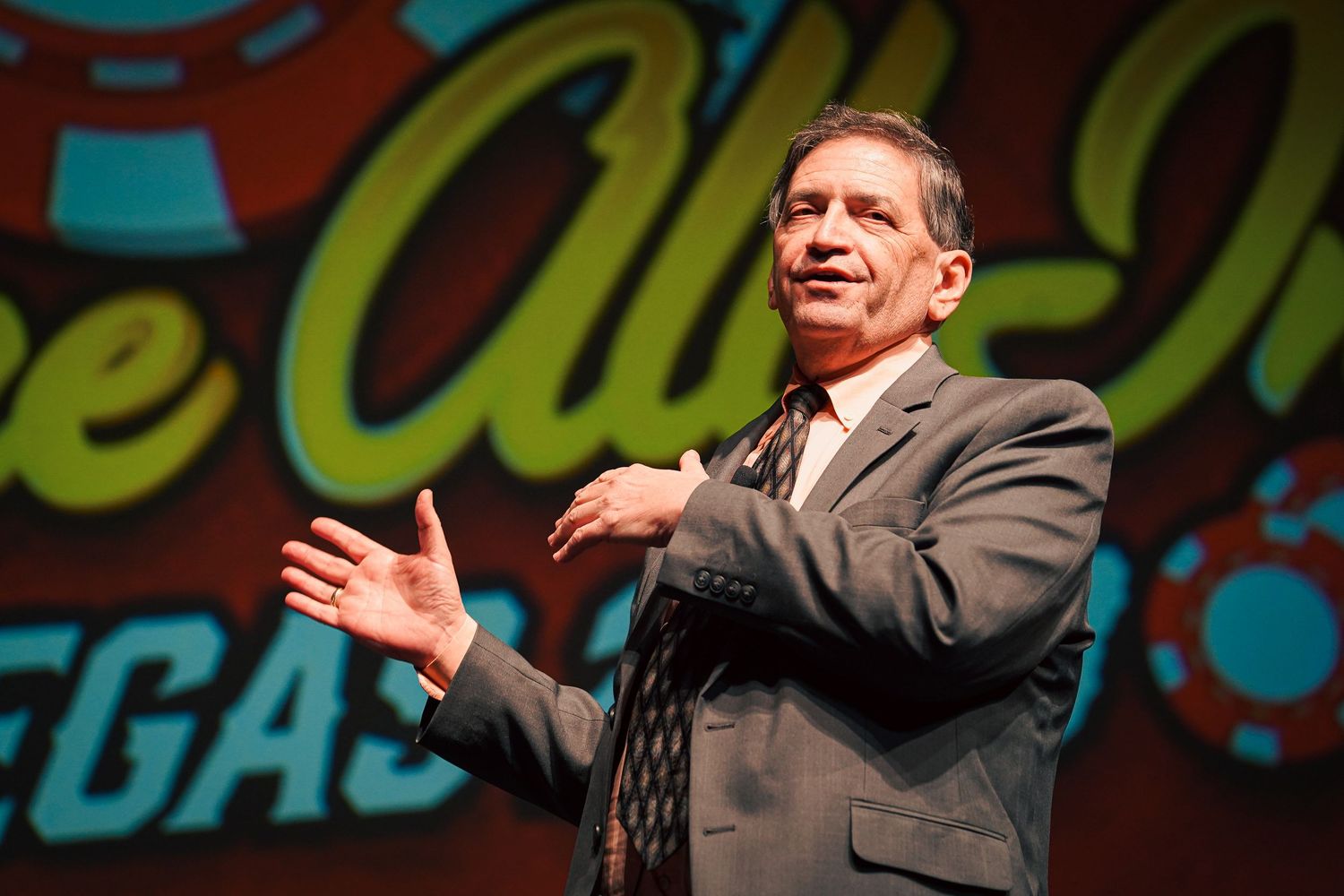 Speaker gestures enthusiastically on stage at Vegas event with colorful backdrop.