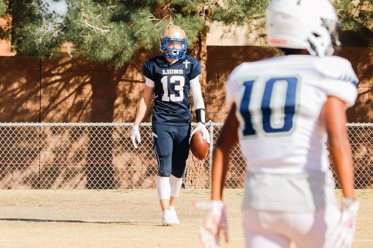 Two opposing football players on field during sunny game with trees in background.