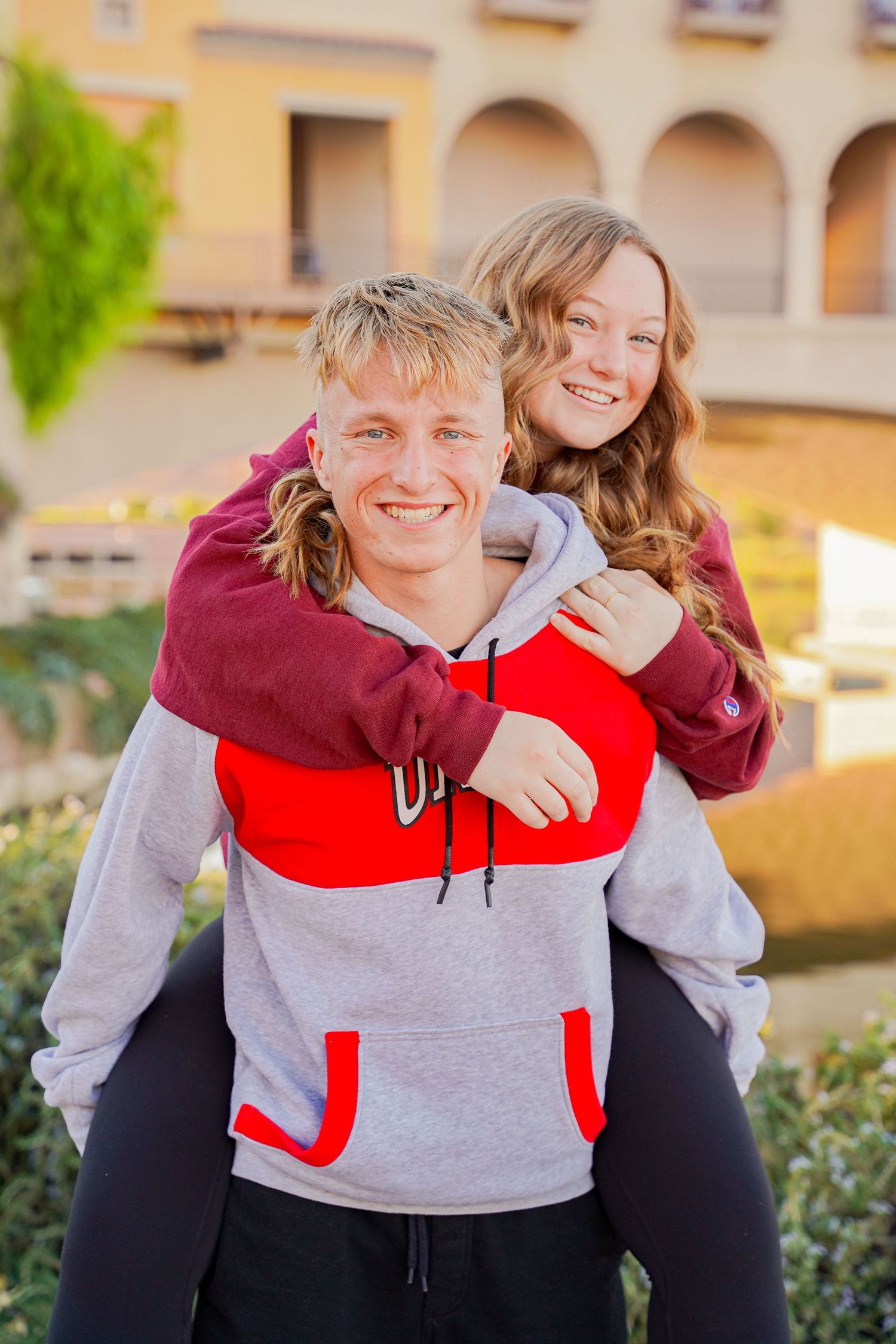 Playful piggyback pose in red and grey sweatshirt against architectural background.