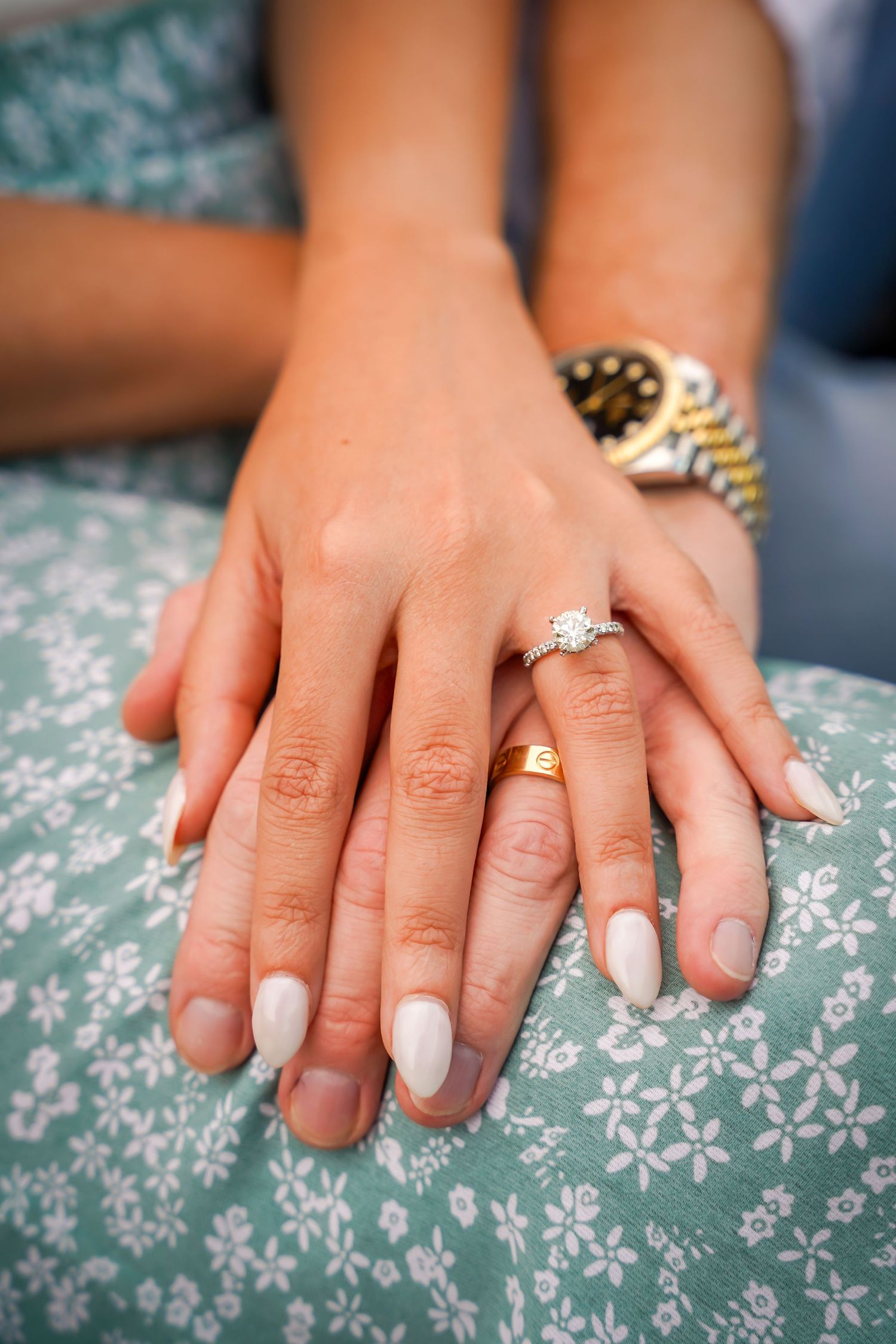 Close up of white manicured hands with diamond engagement ring and gold watch resting on floral pattern fabric.