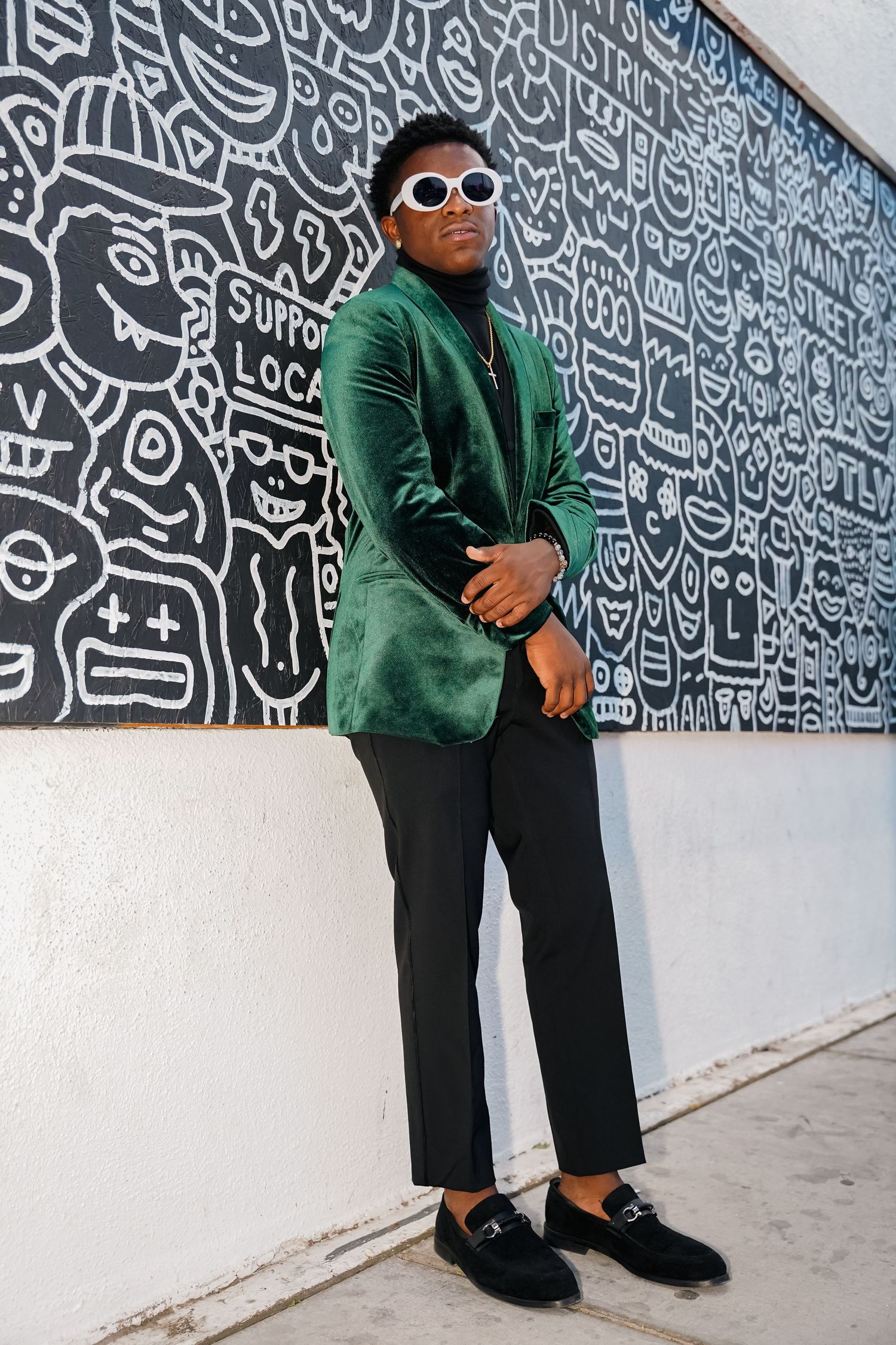 Stylish person wearing green velvet blazer and sunglasses poses against wall with black and white graffiti artwork.