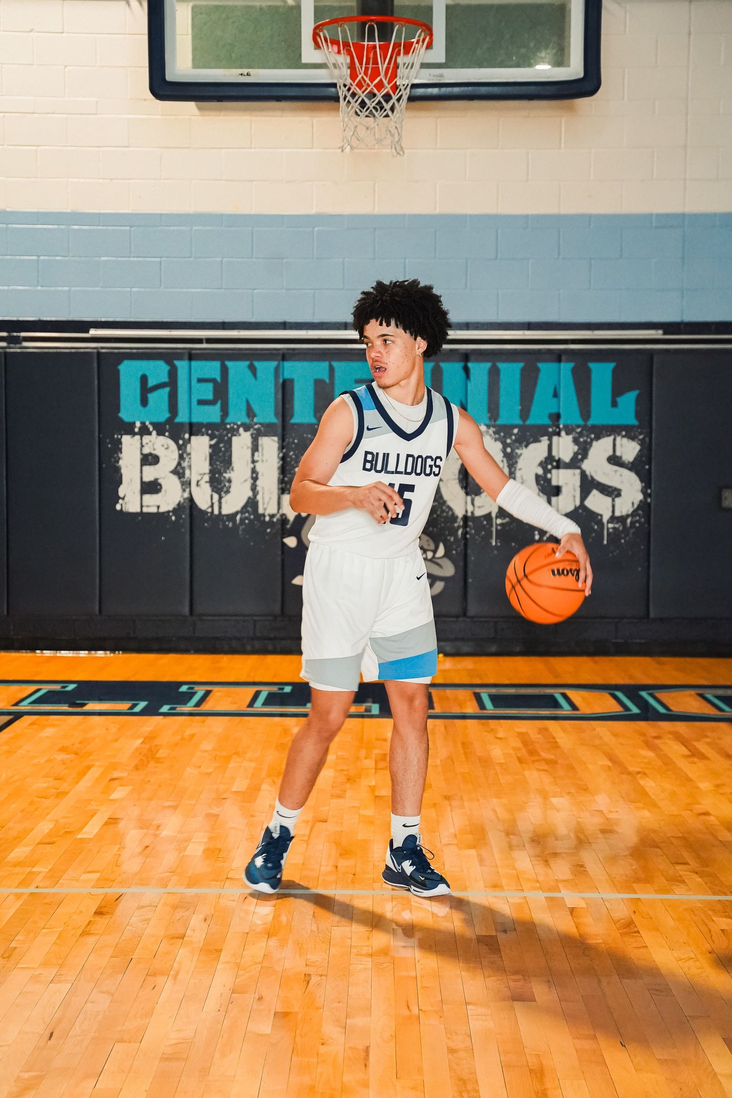 Basketball player dribbles on gymnasium floor with Centennial Bulldogs signage in background.