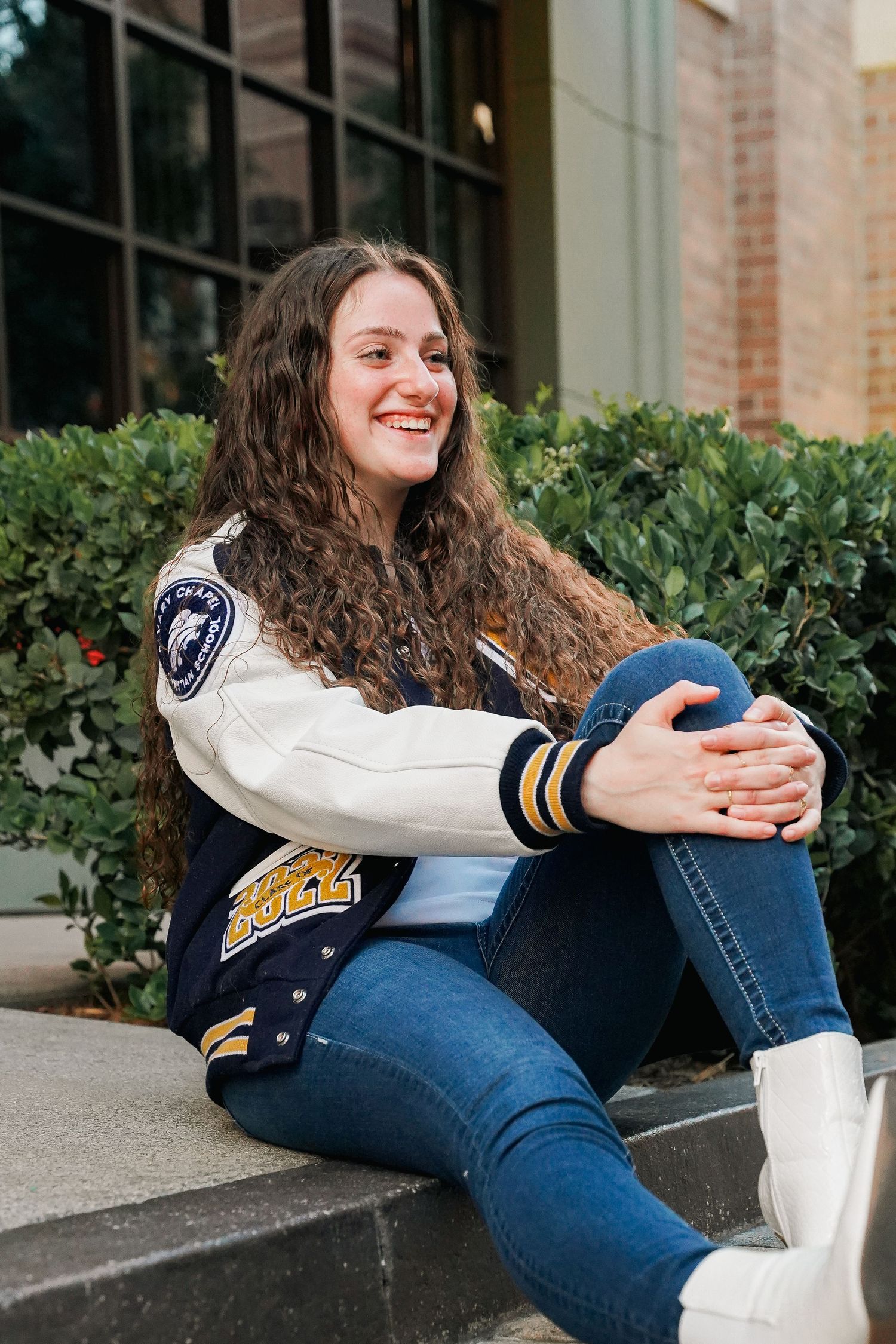 Student in varsity letterman jacket sits on steps outside campus building.