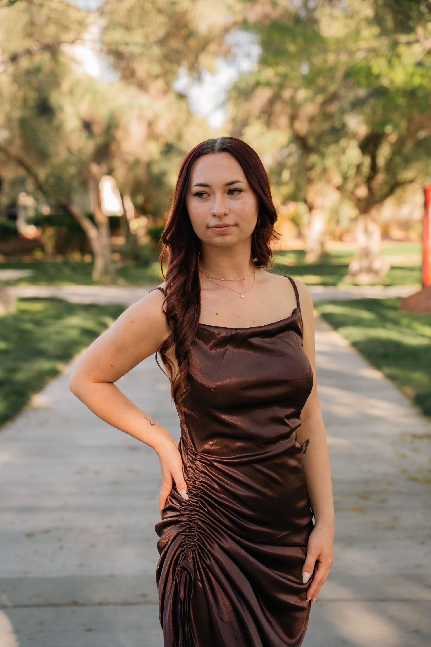 Woman in brown satin dress poses elegantly on garden path with trees.