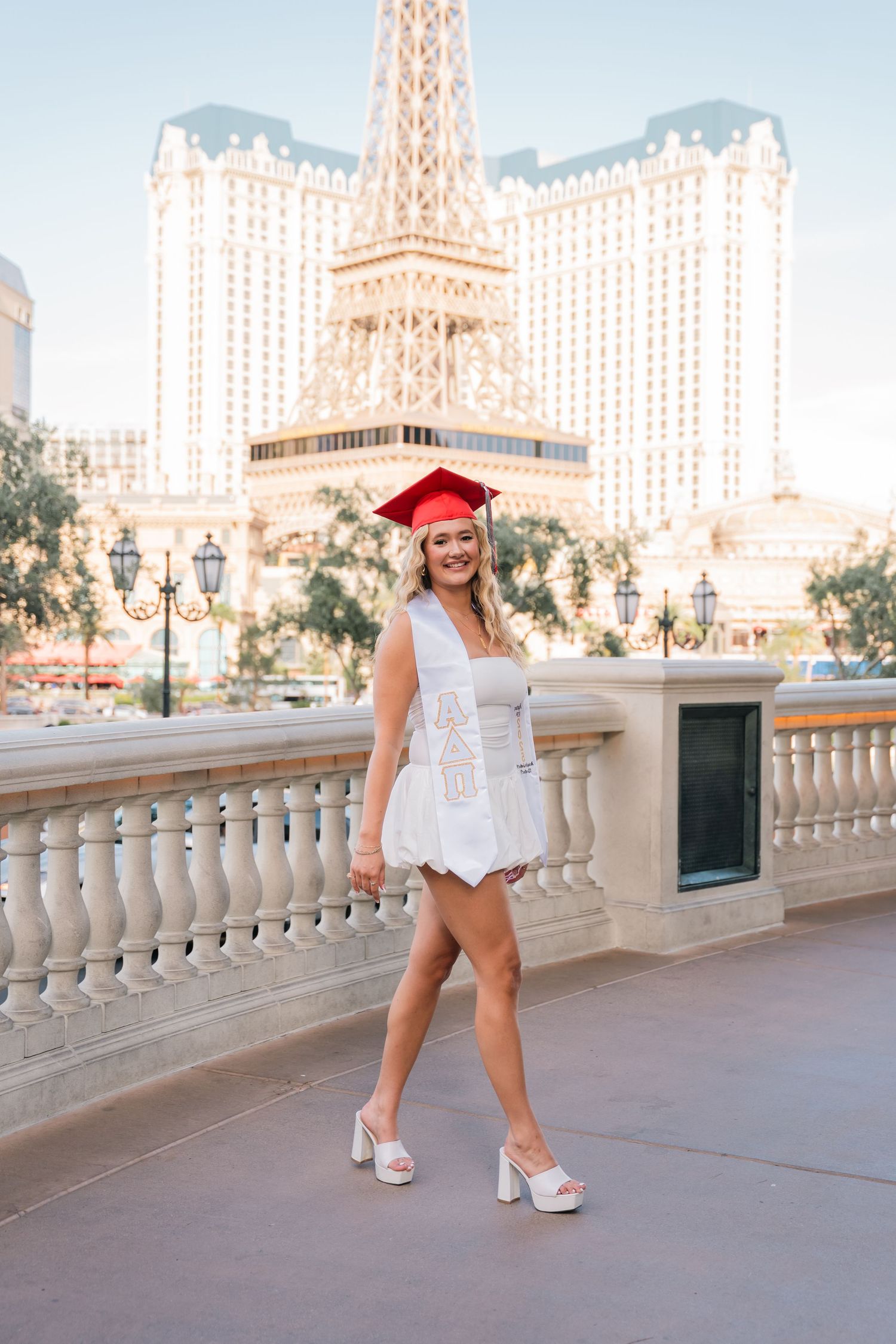 Graduate in white dress and red cap poses on Las Vegas strip with Paris hotel in background.