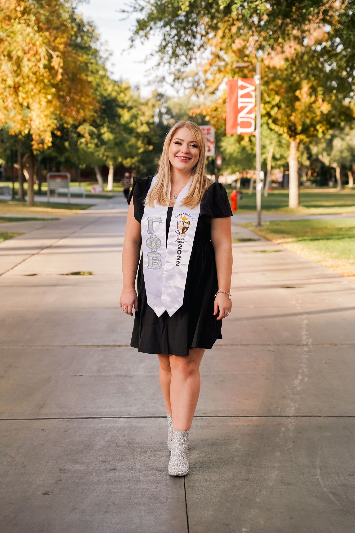 Graduate in black dress and white stoles walks along campus pathway with golden autumn foliage.