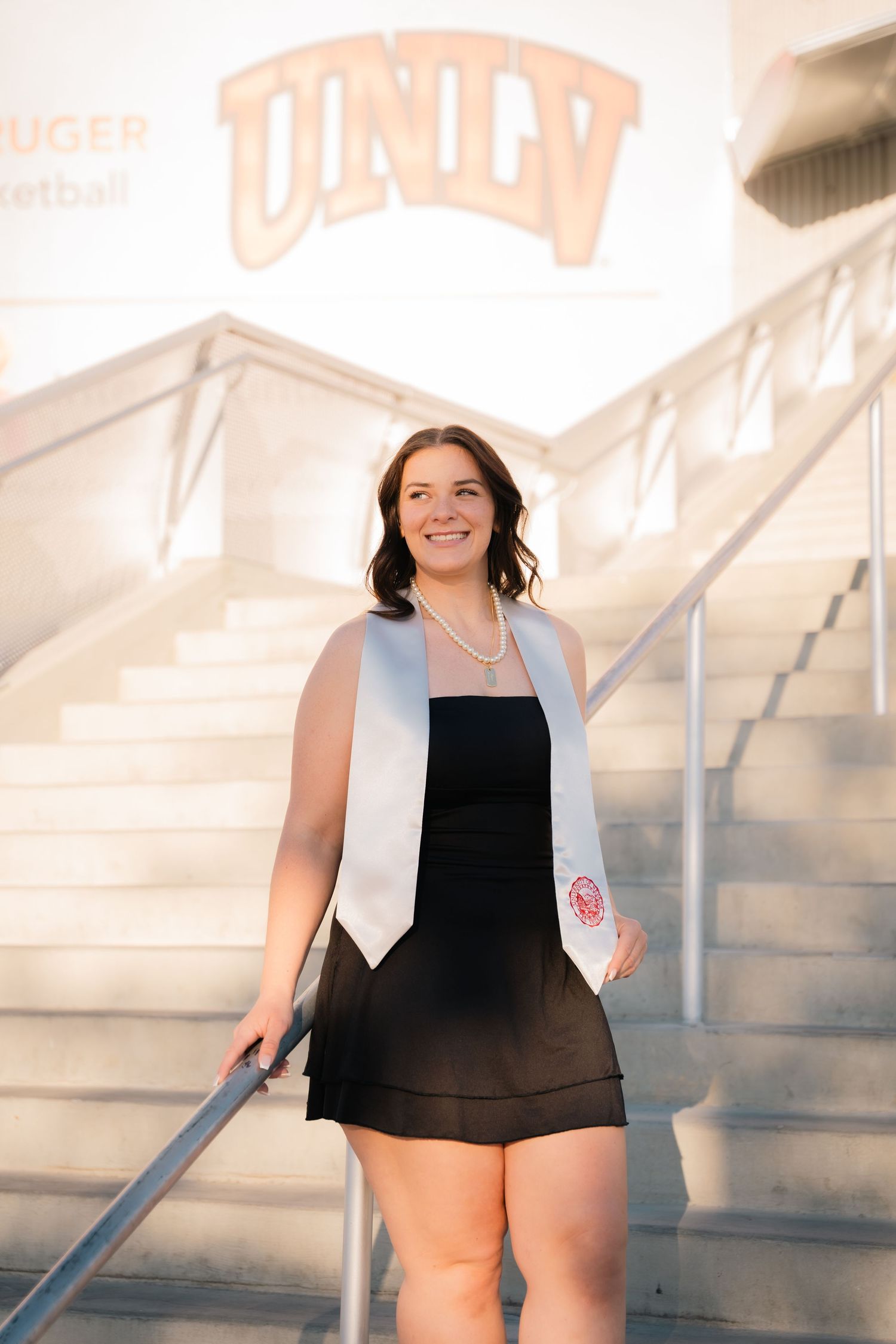 Graduate in black dress and white stole smiles during graduation portrait session on stairs.