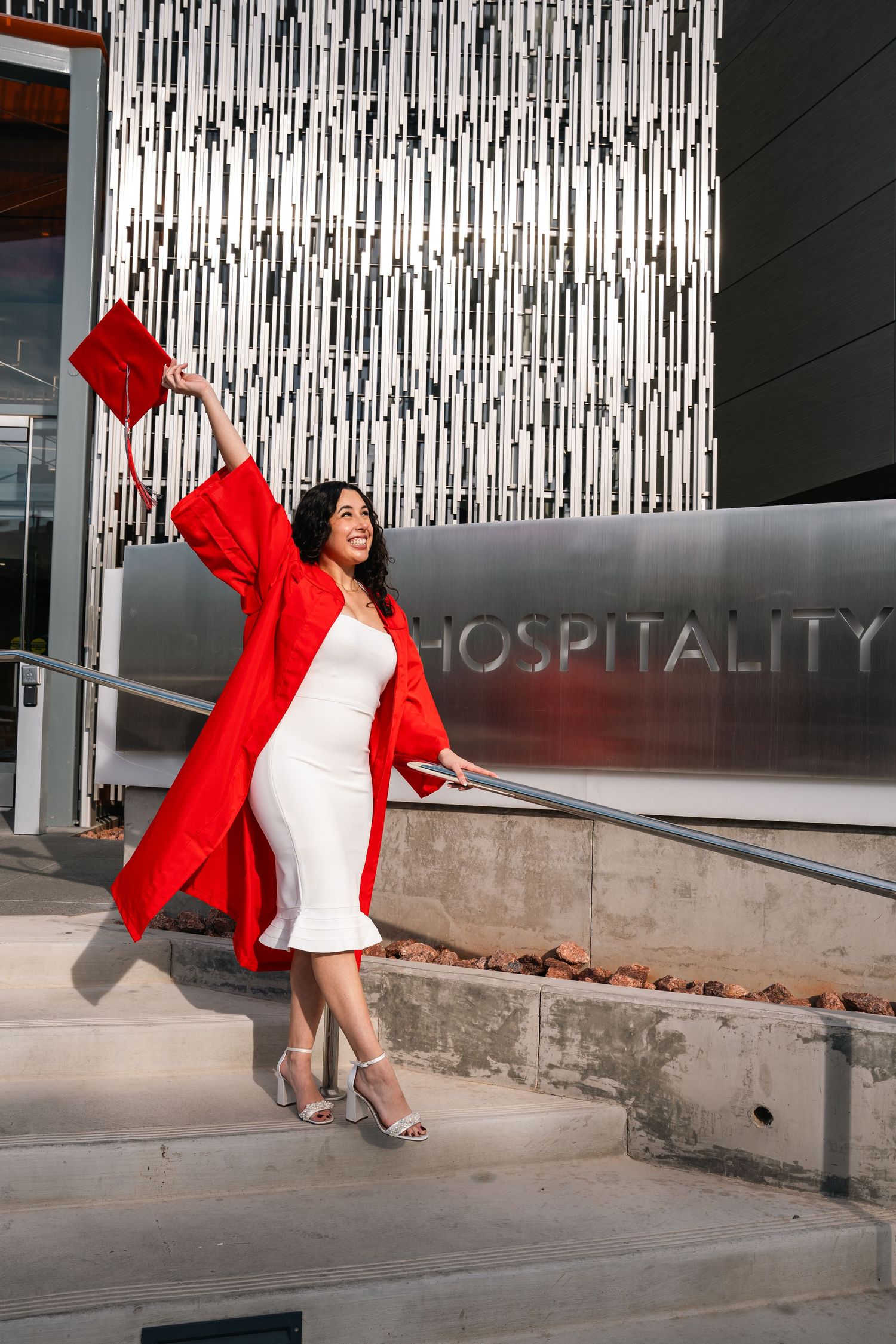 Graduate in white dress and red gown poses at Hospitality building entrance.