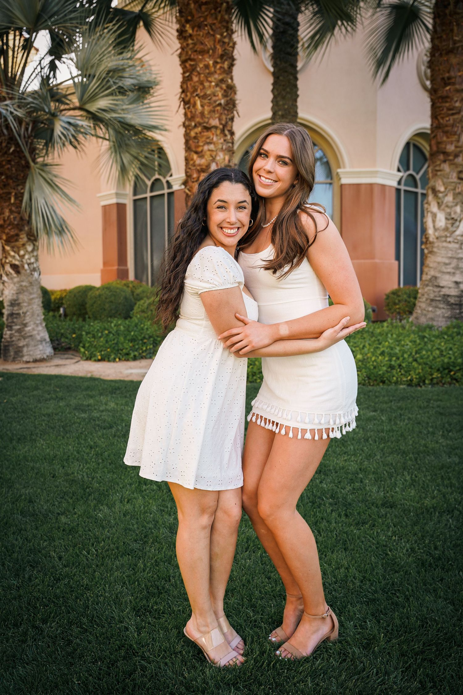 Candid friendship poses in white summer dresses against Mediterranean building and palm trees.