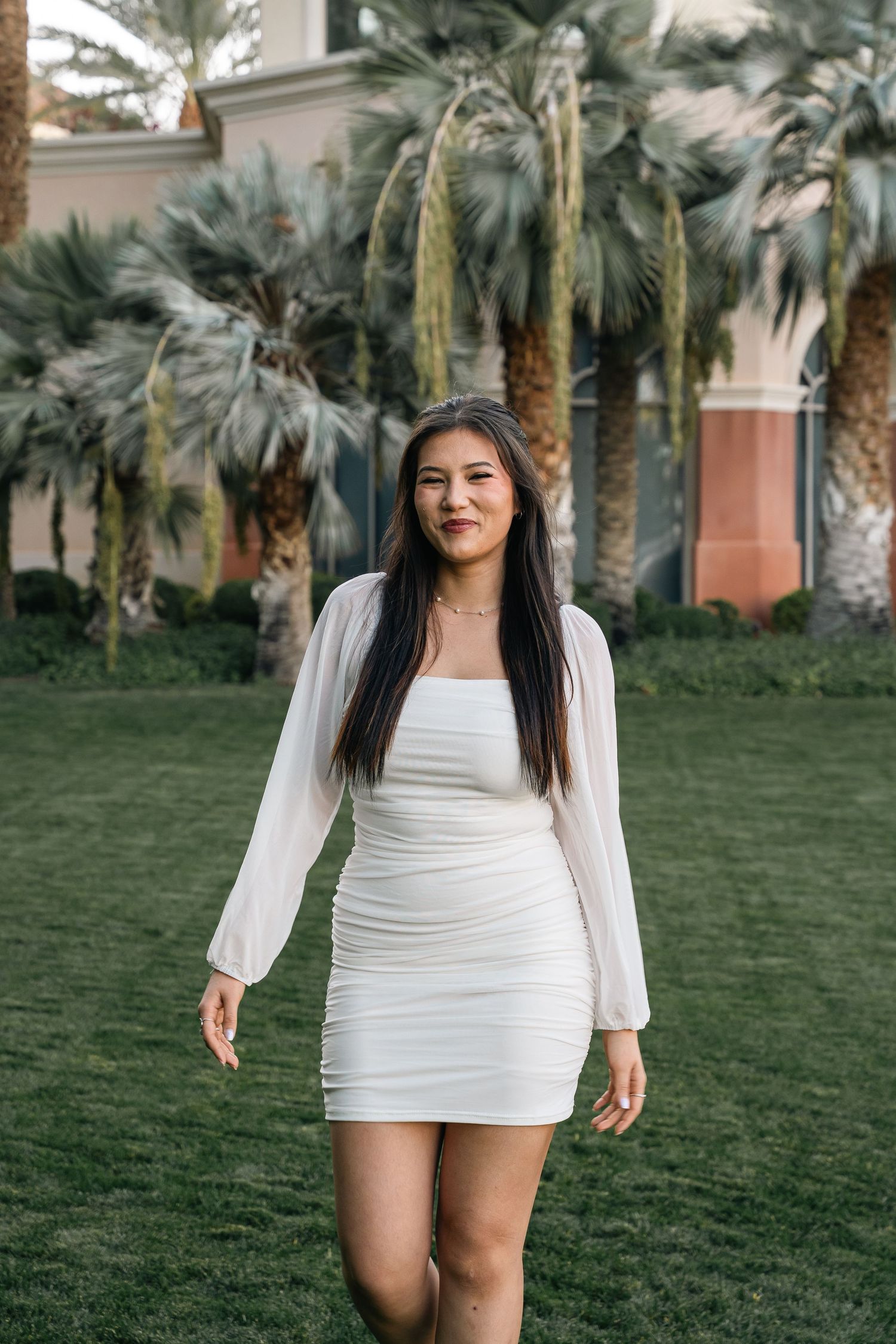 Graduate in white long-sleeved dress poses on lawn with palm trees and Spanish architecture.