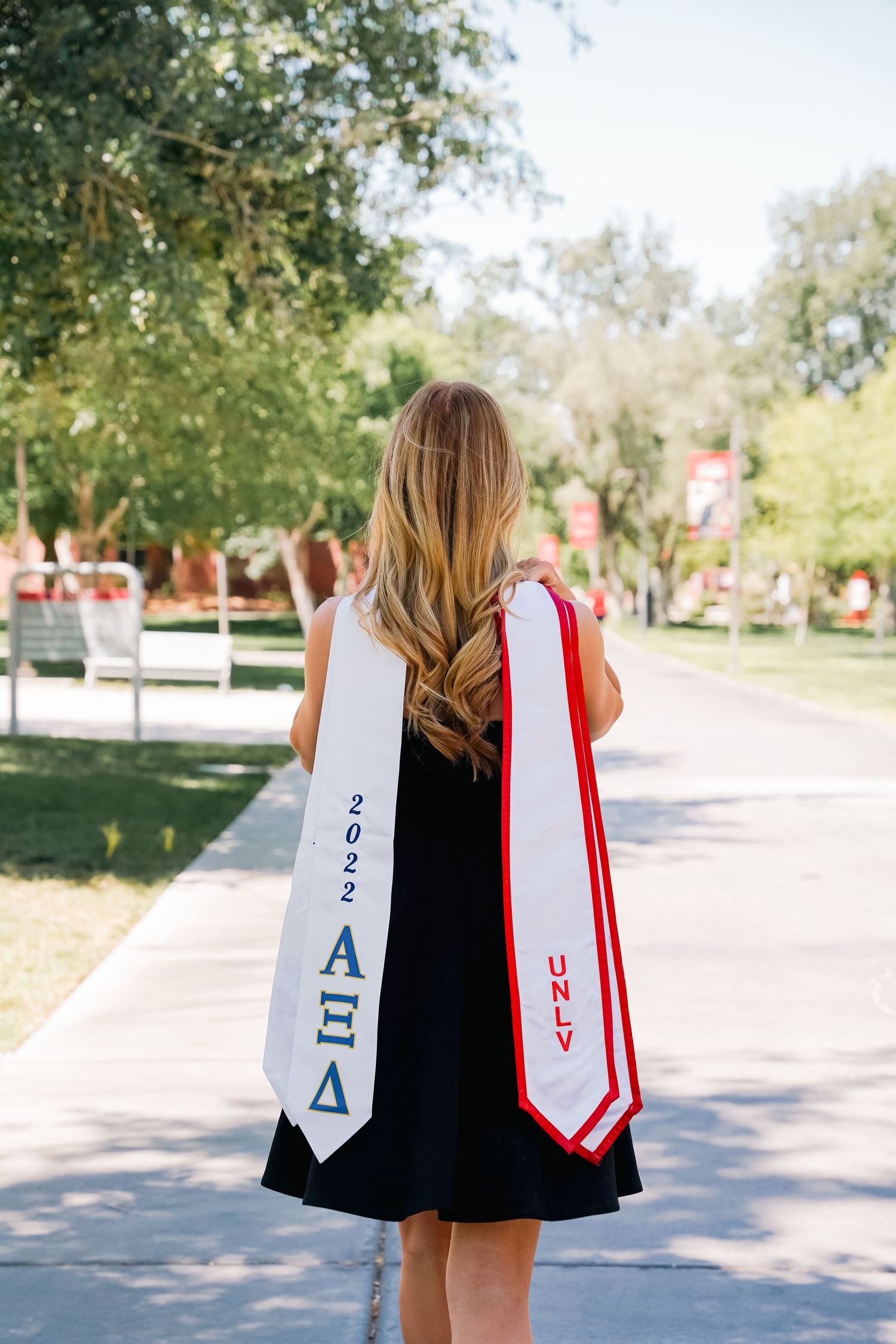 Graduate wearing black dress displays sorority graduation stoles while walking on campus pathway.
