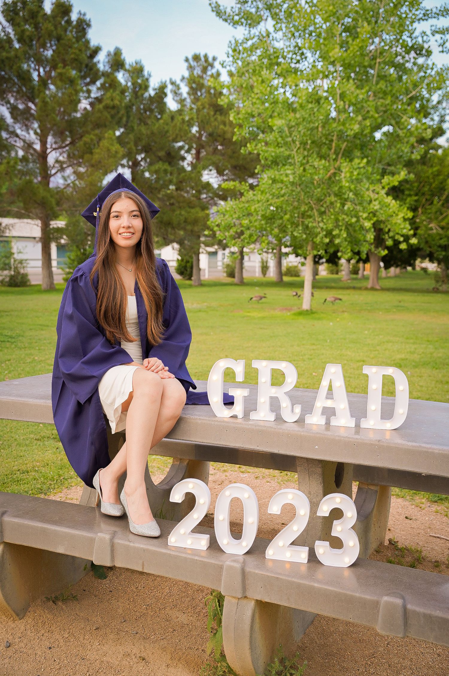 Graduate in blue cap and gown poses with GRAD 2023 letters on outdoor bench.