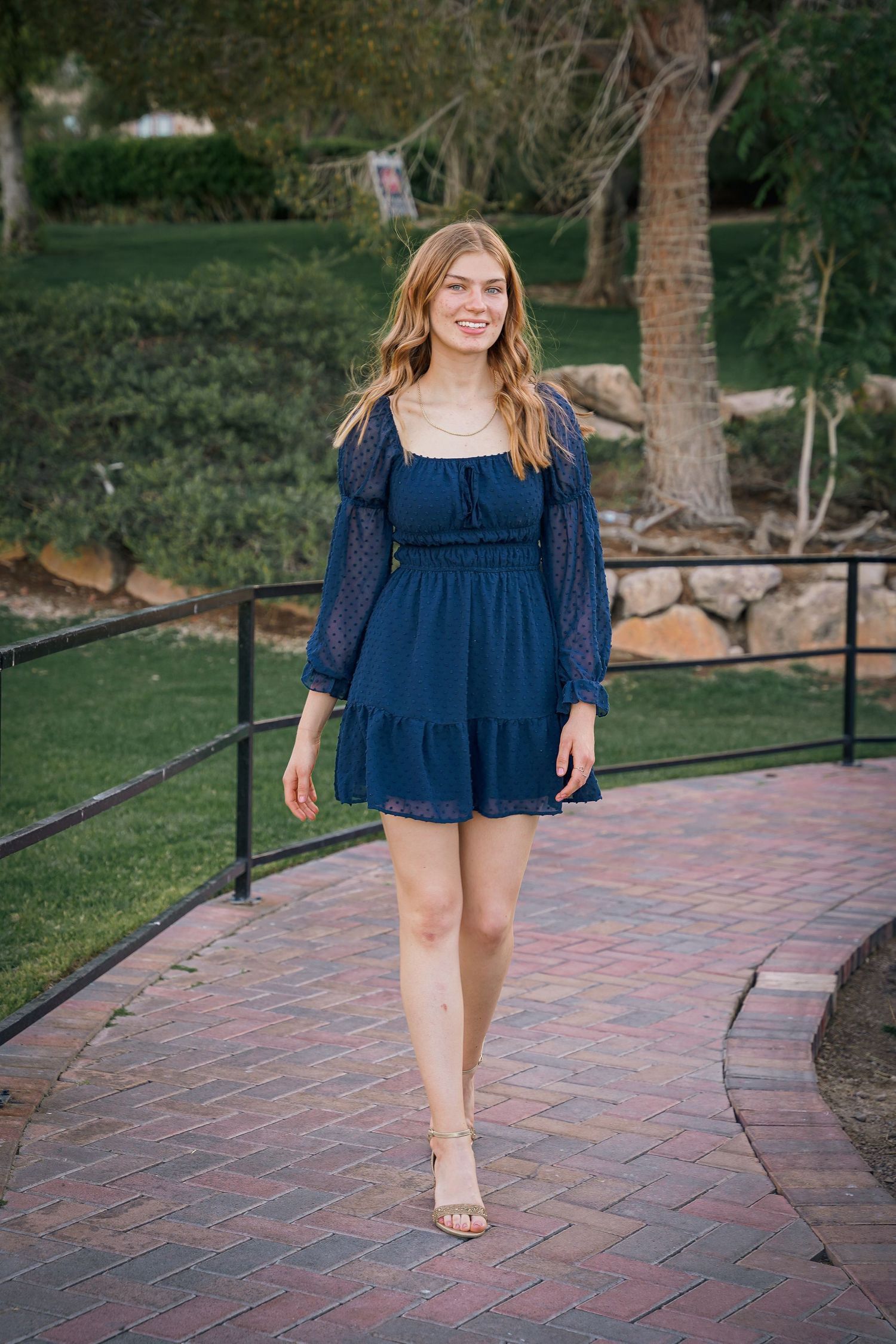 Woman in navy blue summer dress walking along a curved brick pathway in a garden setting.