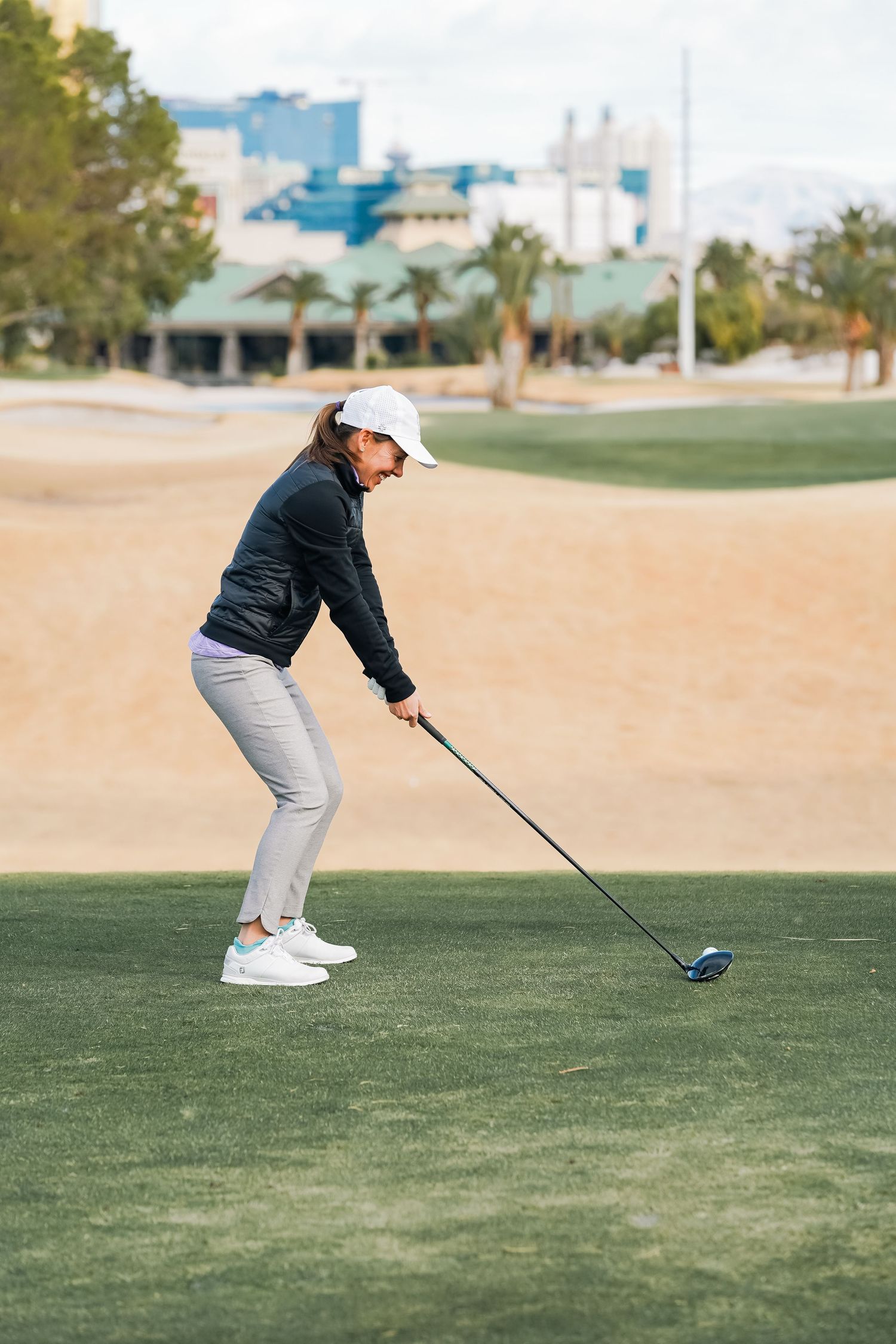 Golfer sets up for a drive on desert golf course with palm trees and resort buildings visible.