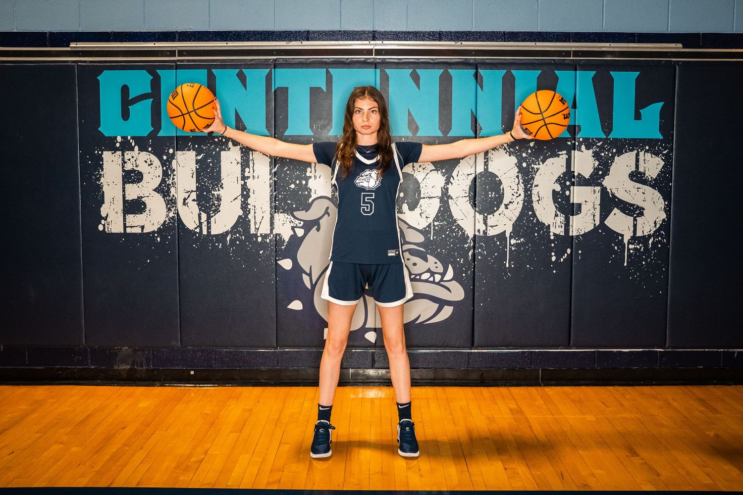 Basketball player practicing stance drills on wooden court with school branding visible.