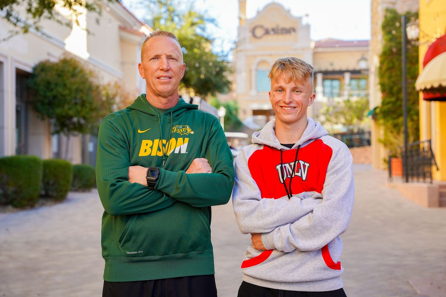 Two people wearing athletic sweatshirts displaying NDSU Bison and UND logos pose on a sunny campus path.