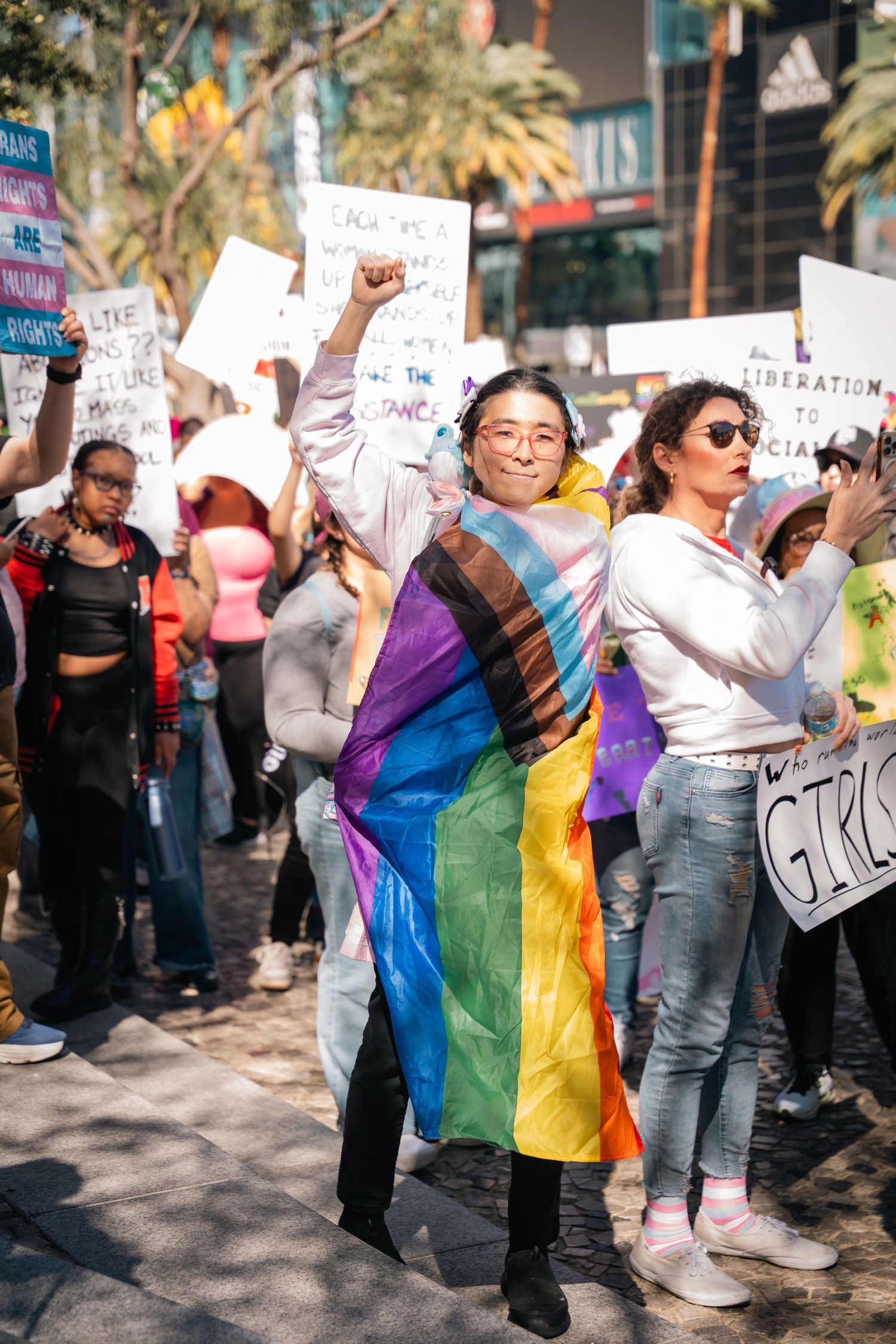 Protesters gather at a rally with rainbow pride flags and signs demonstrating for equality and rights.