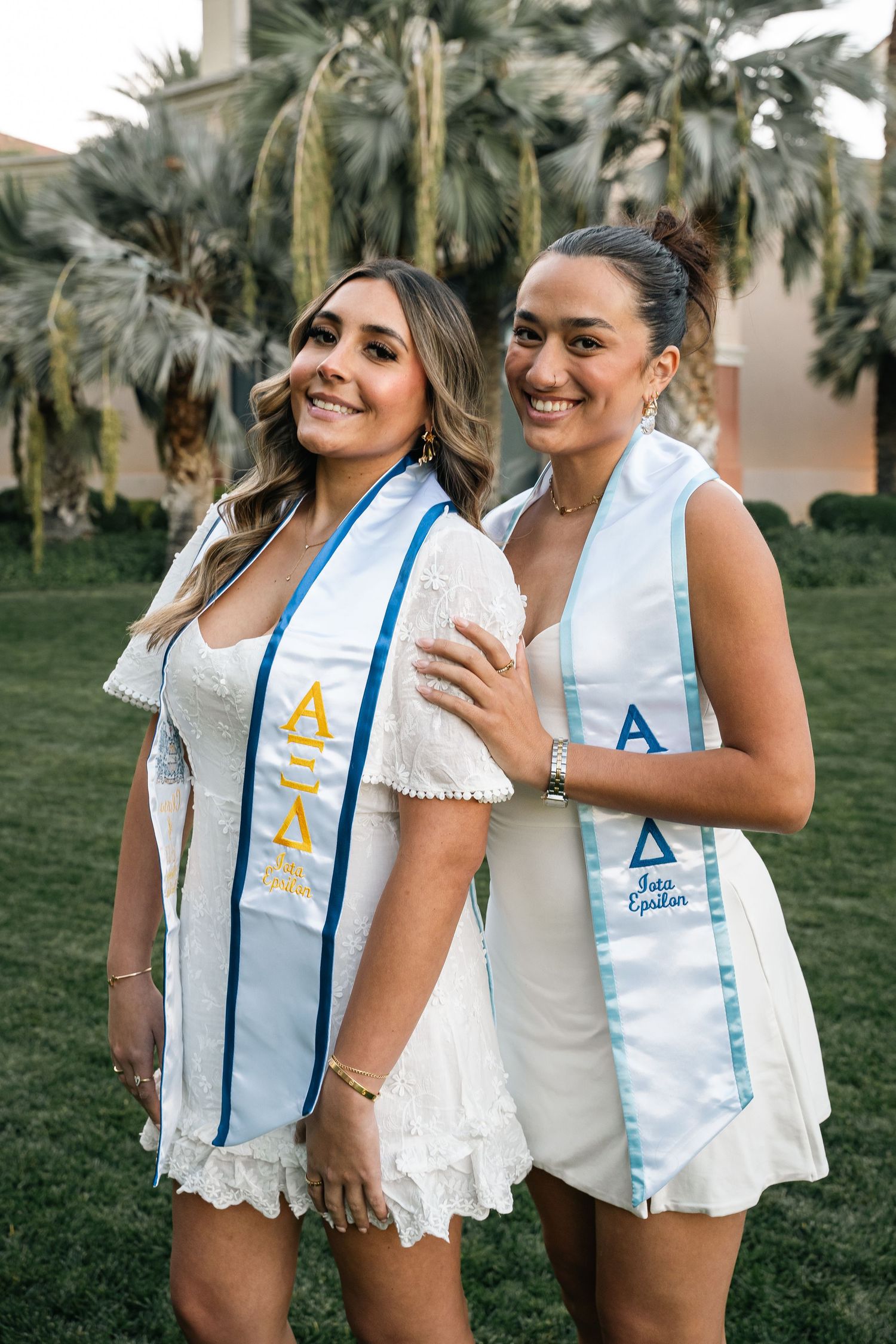 Two students wearing white dresses and light blue graduation sashes pose together outdoors.