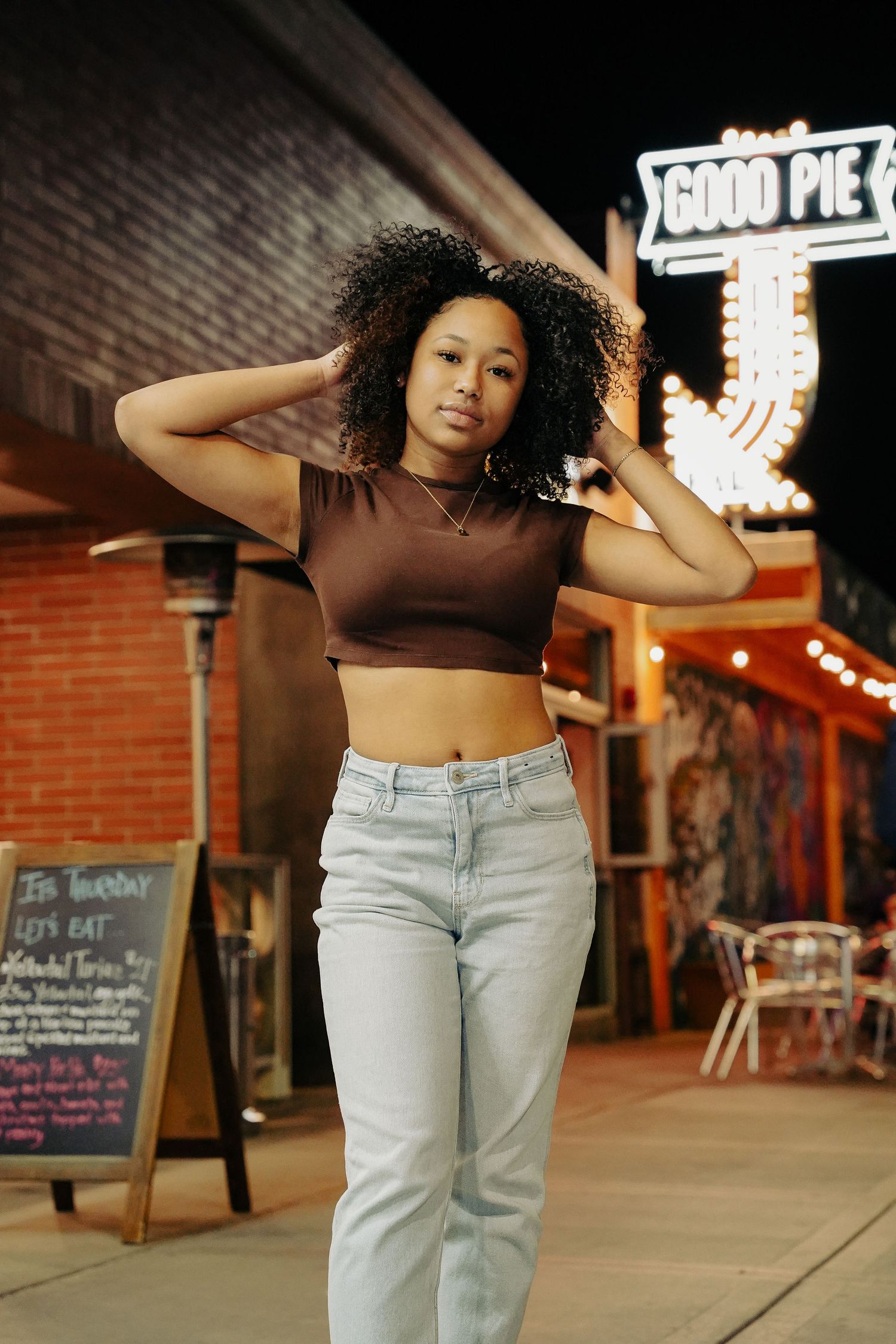Woman in brown crop top and light jeans poses outside Good Pie restaurant at night.
