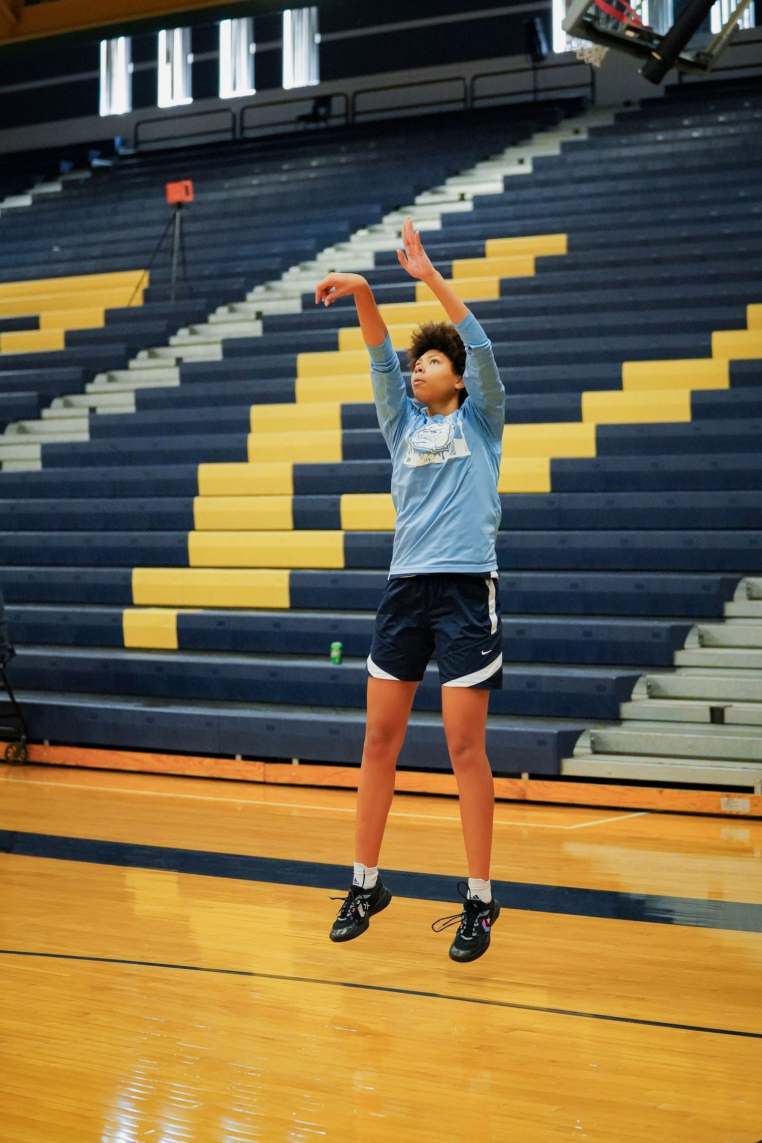 Player practices shooting form in empty gymnasium with blue and yellow bleachers.