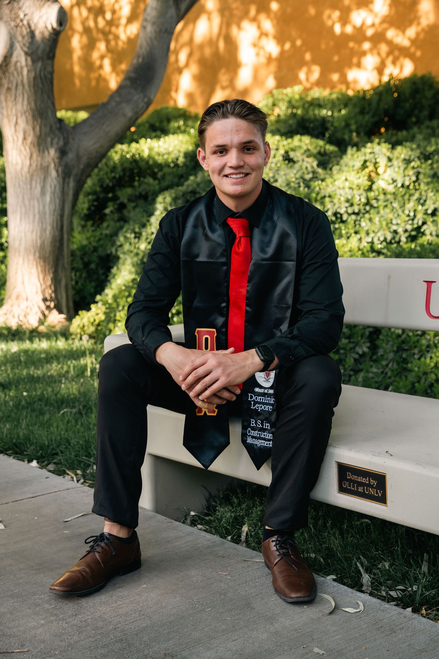 Graduate in formal attire poses on campus bench wearing graduation stole.