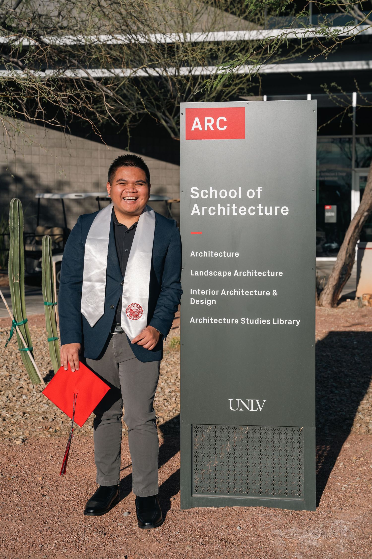 Graduate poses next to UNLV School of Architecture sign in formal attire.