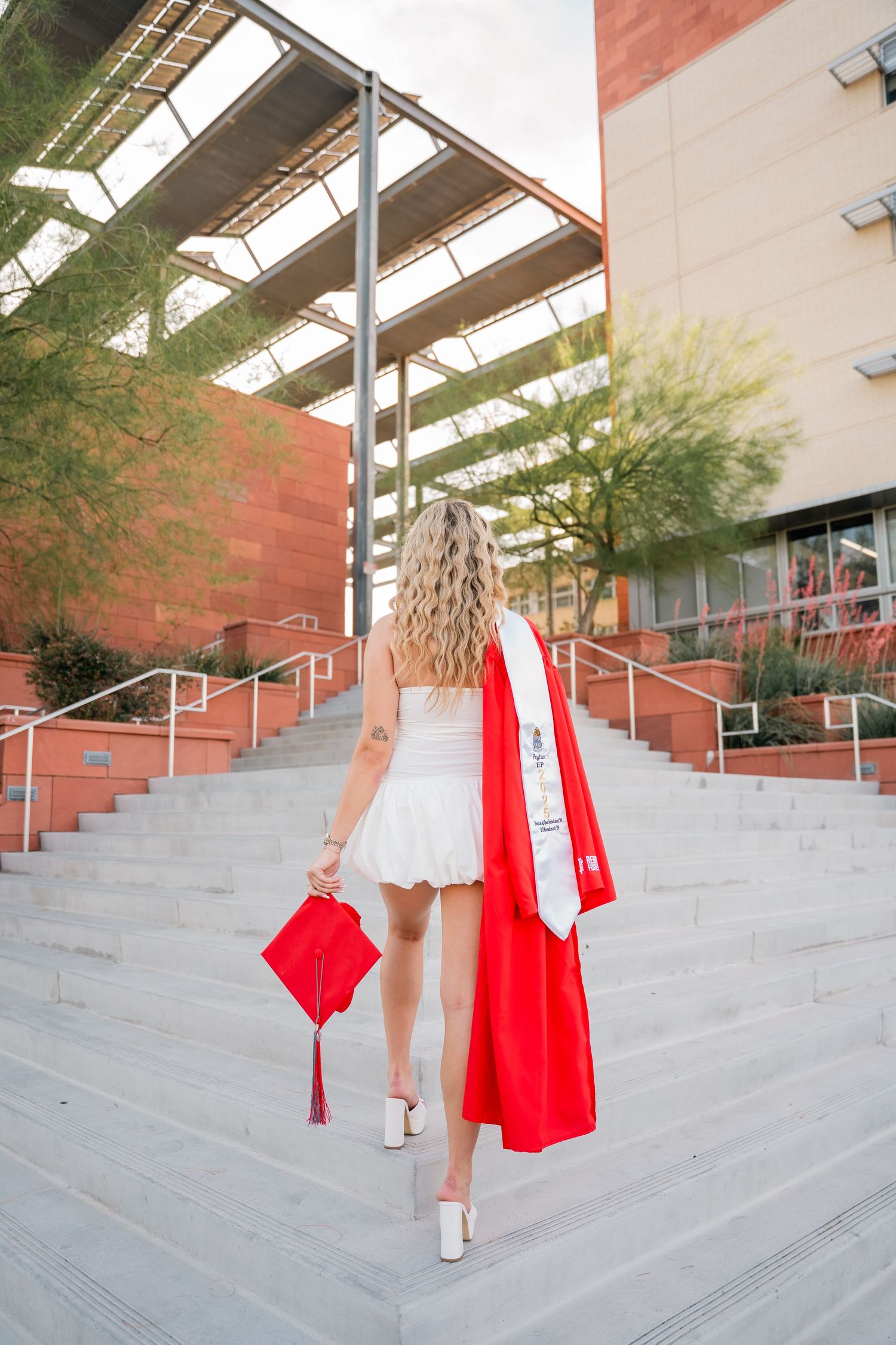 Graduate in red cap and white dress poses on concrete campus steps.