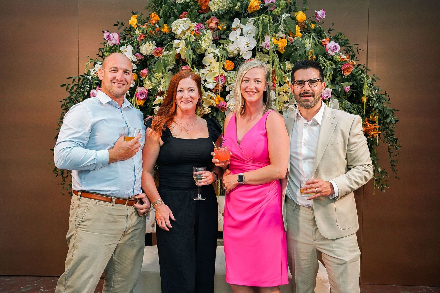 Four guests pose in front of a lush floral arch during a celebratory event.